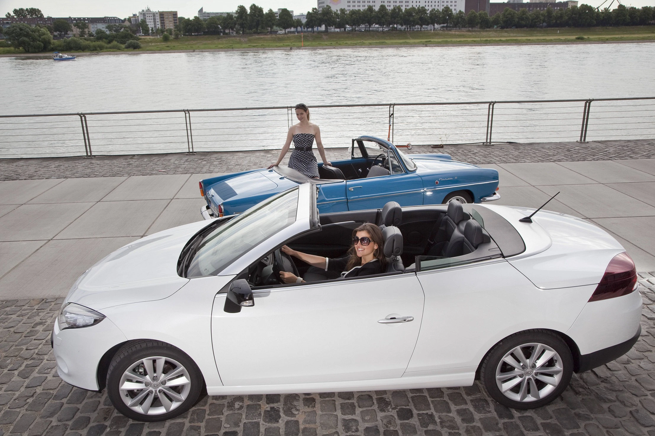 Zwei Frauen stehen und sitzen in Cabrios an einer Uferpromenade, mit einem Fluss und Bäumen im Hintergrund.