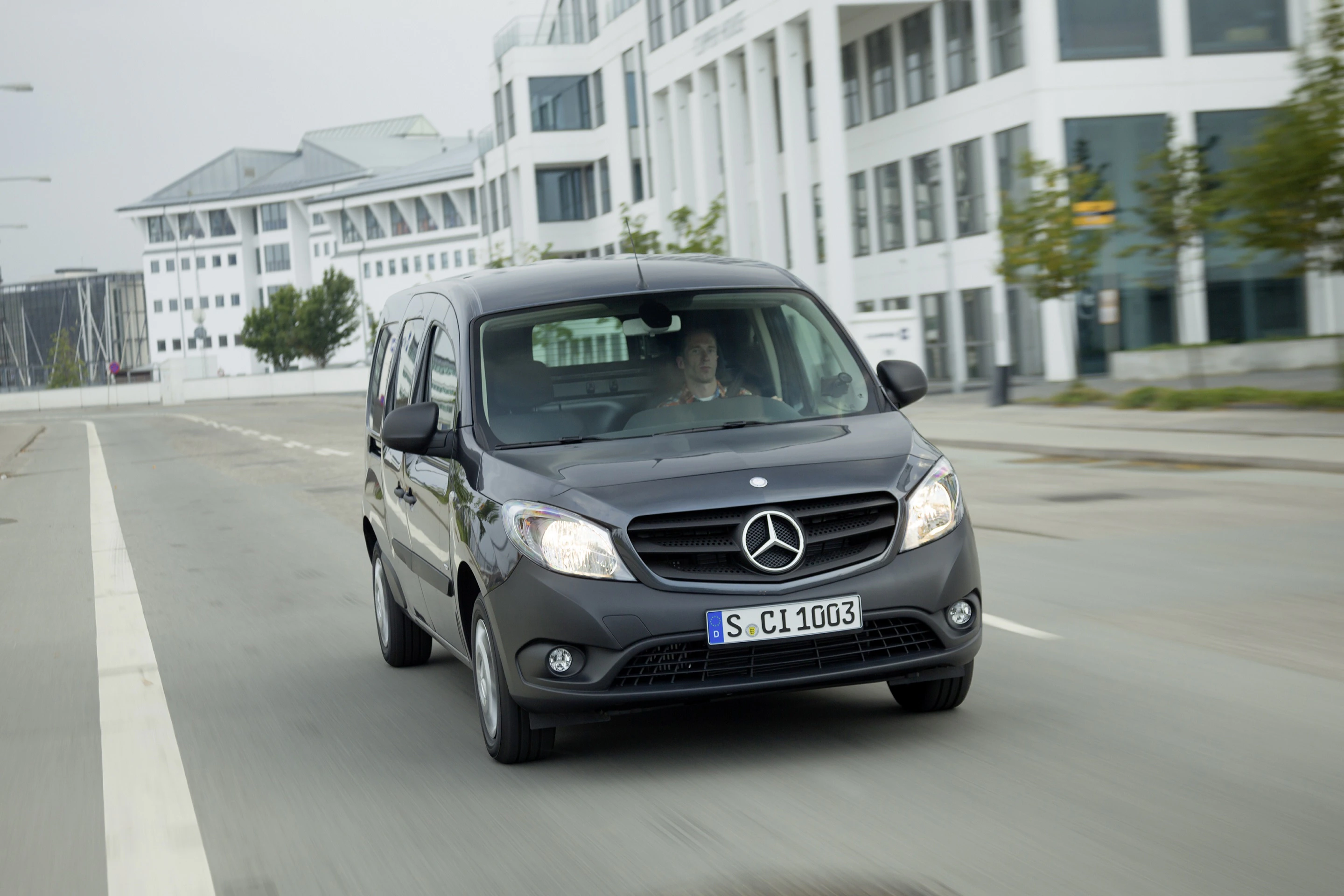 Grauer Mercedes-Benz Citan fährt auf einer leeren Stadtstraße, umgeben von modernen Gebäuden. Ein Mann sitzt am Steuer.