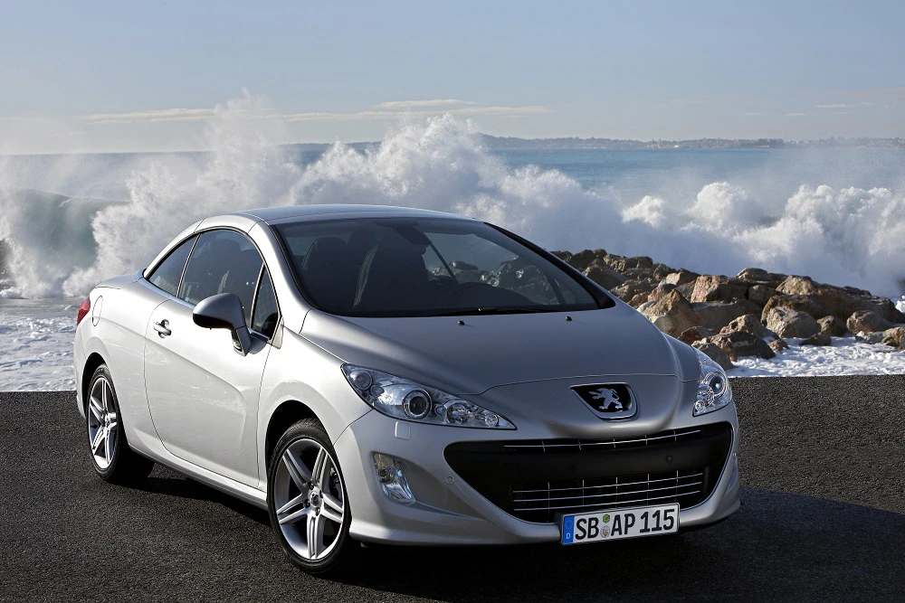 Silberner Peugeot 308 CC parkt auf Asphalt nahe des Meeres, hohe Wellen im Hintergrund. Ruhige, sonnige Küstenszene.