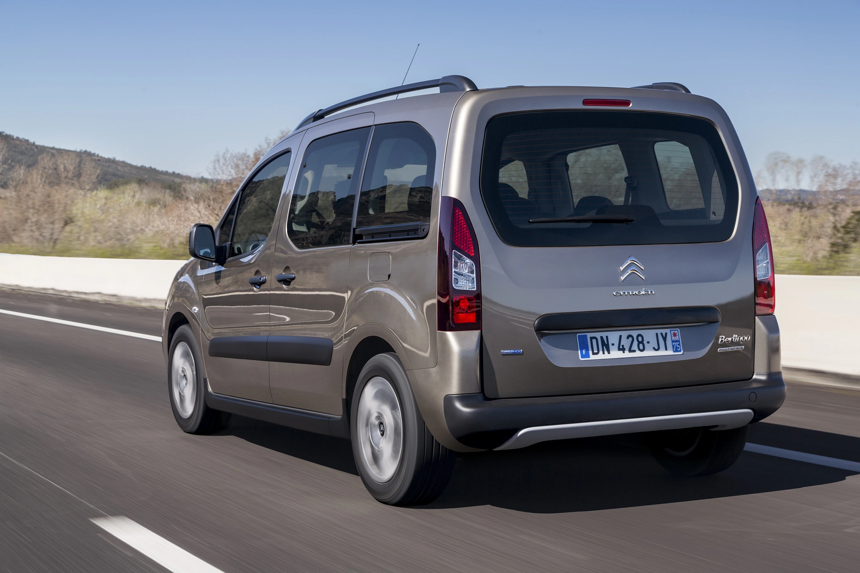 Brauner Citroën Berlingo fährt auf einer Landstraße. Hinten und seitlich zu sehen, mit Bäumen im Hintergrund.