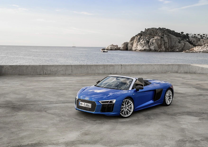 Blauer Sportwagen Cabrio auf engem Parkplatz an der Küste mit Felsen und Meer im Hintergrund.