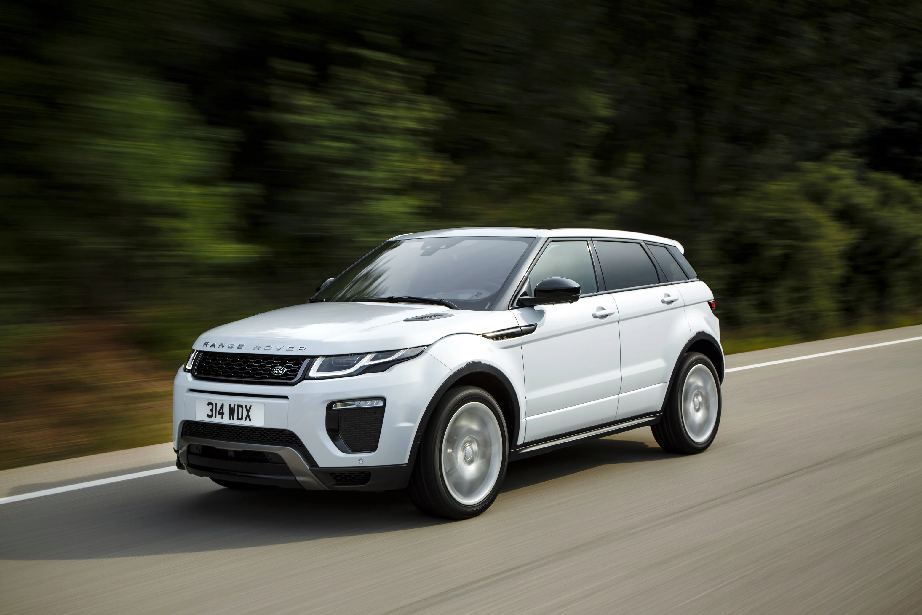 Weißer SUV fährt auf einer Straße durch eine grüne, bewaldete Landschaft. Fahrzeug ist dynamisch und modernes Design.