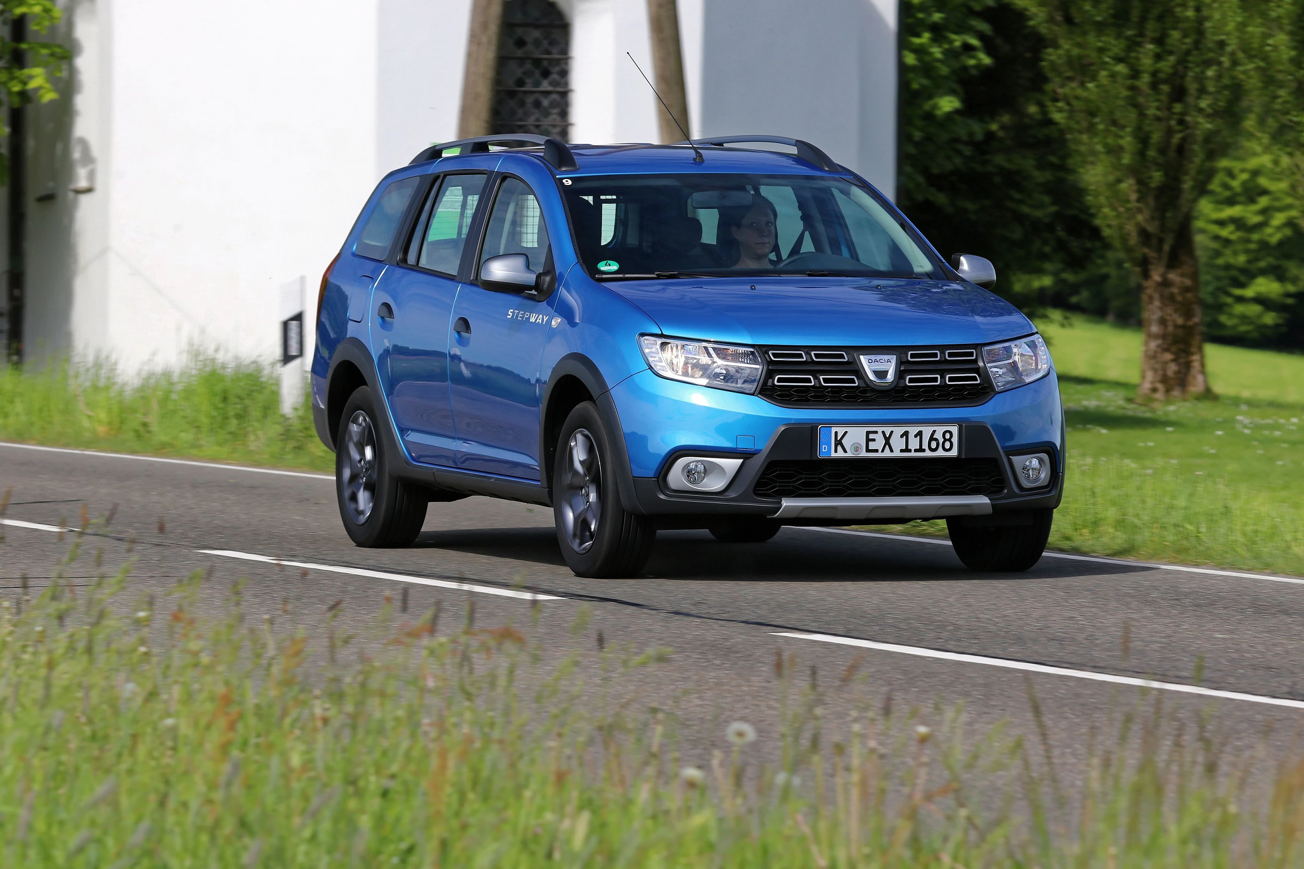 Blauer SUV fährt auf Landstraße, umgeben von Wiesen und Bäumen, Kirche im Hintergrund. Fahrzeugnummer: K-EX1168.