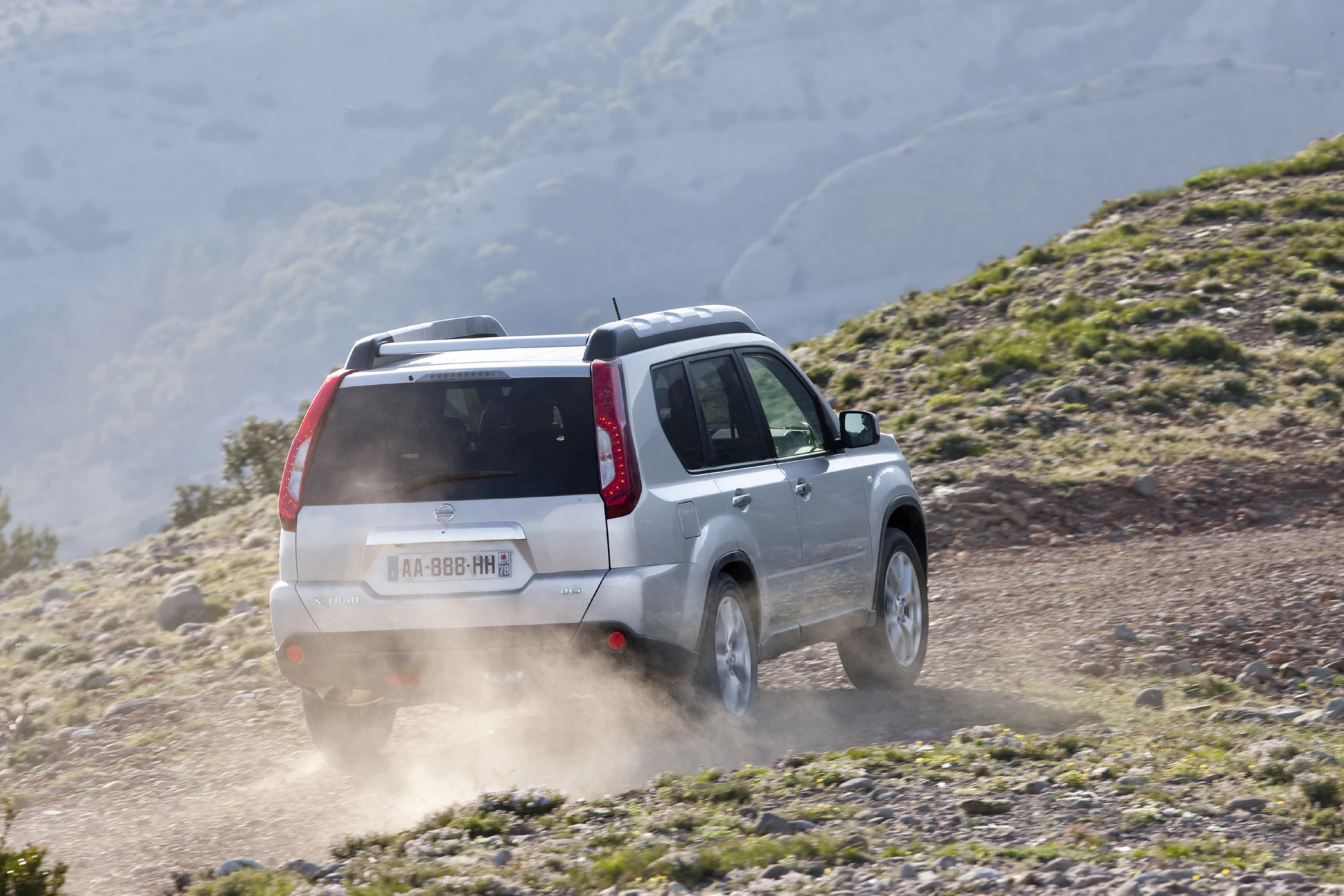 Silberner SUV fährt auf einem staubigen Weg durch eine hügelige Landschaft. Im Hintergrund sind Berge und Bäume zu sehen.
