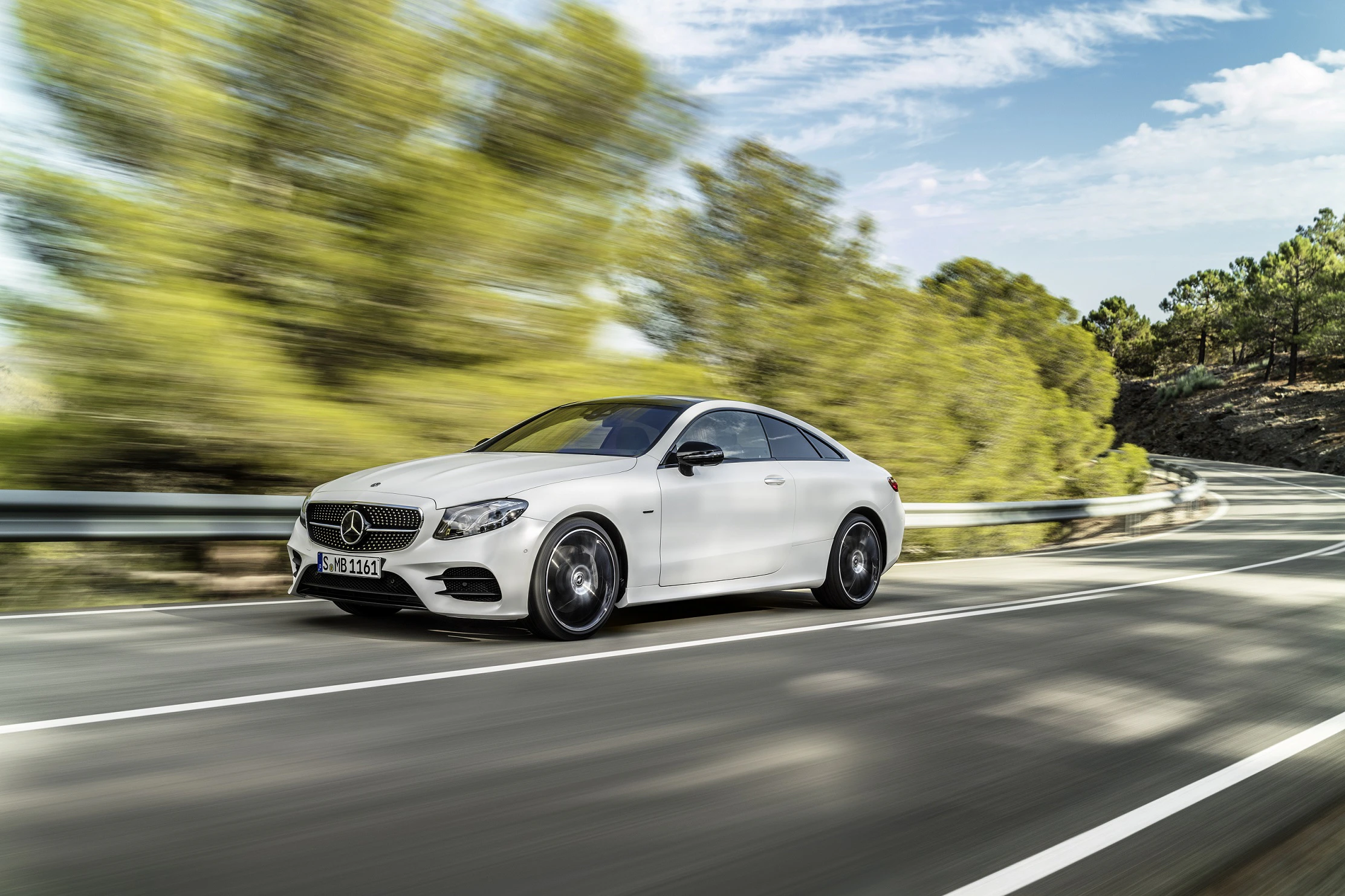 Ein weißes Mercedes-Benz Coupé fährt schnell auf einer kurvigen Landstraße, umgeben von Bäumen und bewölktem Himmel.