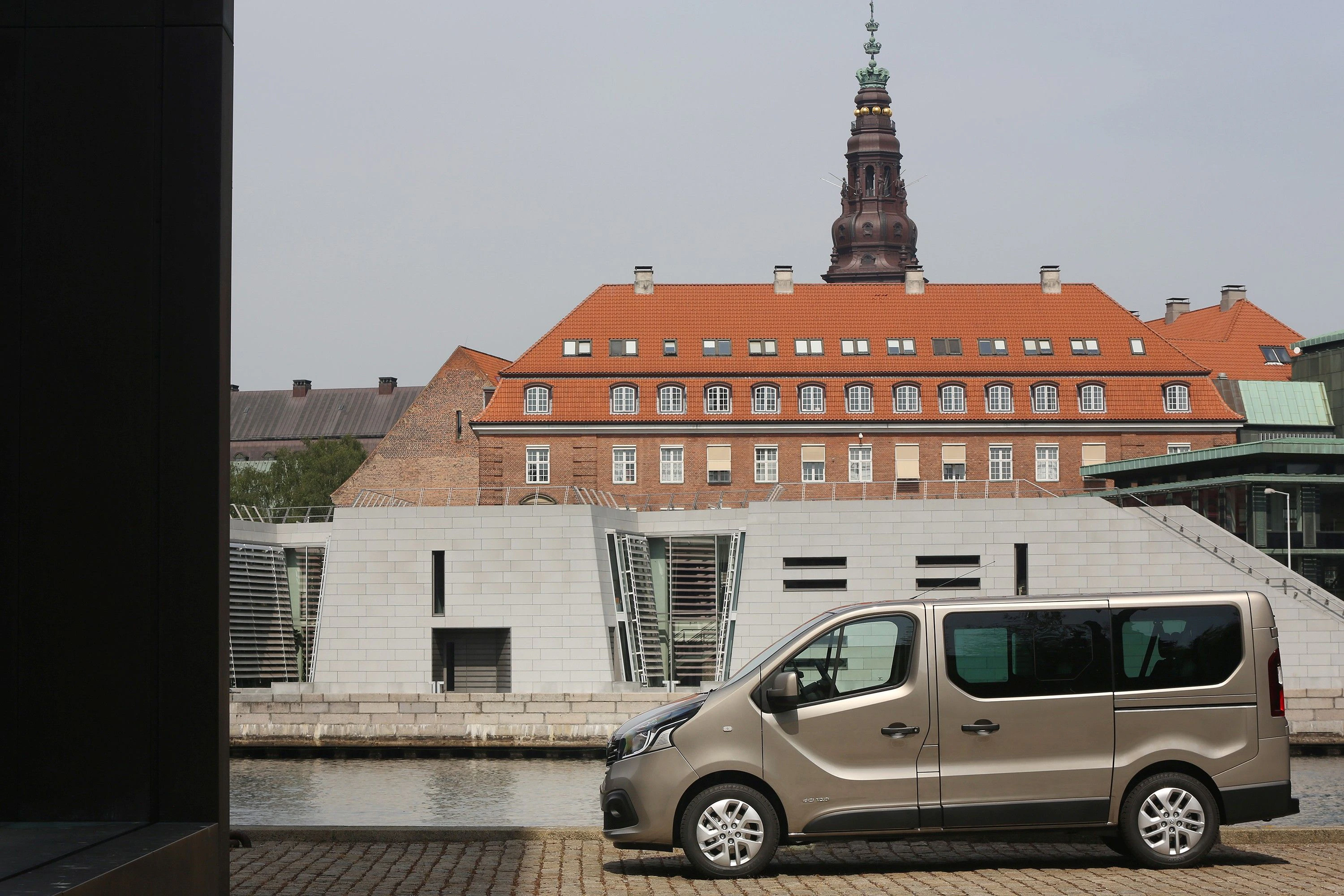 Ein beige Kleinbus steht vor einem modernen Gebäude mit Wasserfront; im Hintergrund ein historisches Gebäude mit rotem Ziegeldach und Turm.