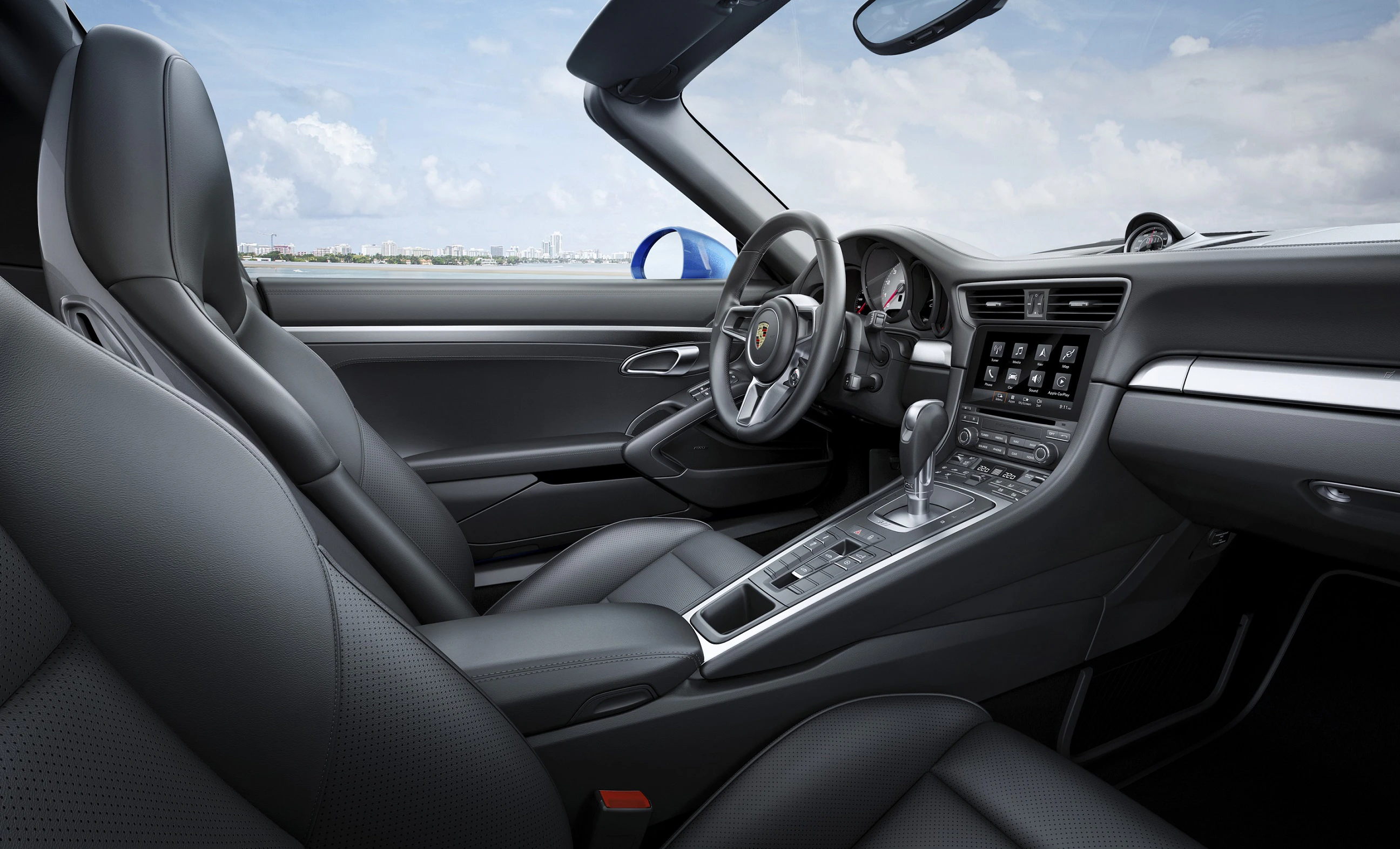Innenraum eines modernen Cabriolets mit Ledersitzen, Sportlenkrad, Automatikgetriebe und Multimedia-Bildschirm, unter wolkigem Himmel.