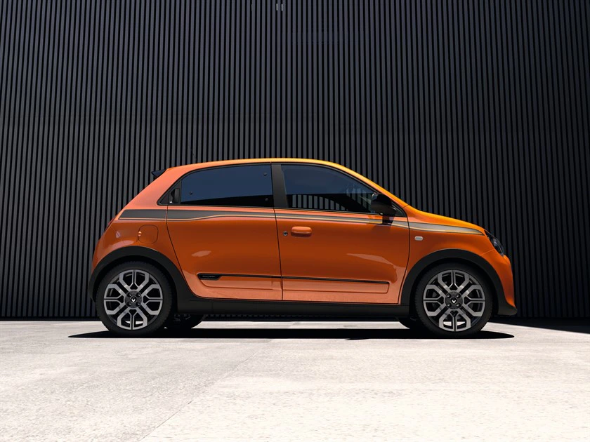 Orangefarbener Kleinwagen vor einer schwarzen, gerillten Wand. Das Auto steht seitlich, zeigt modernes Design und stilvolle Felgen.
