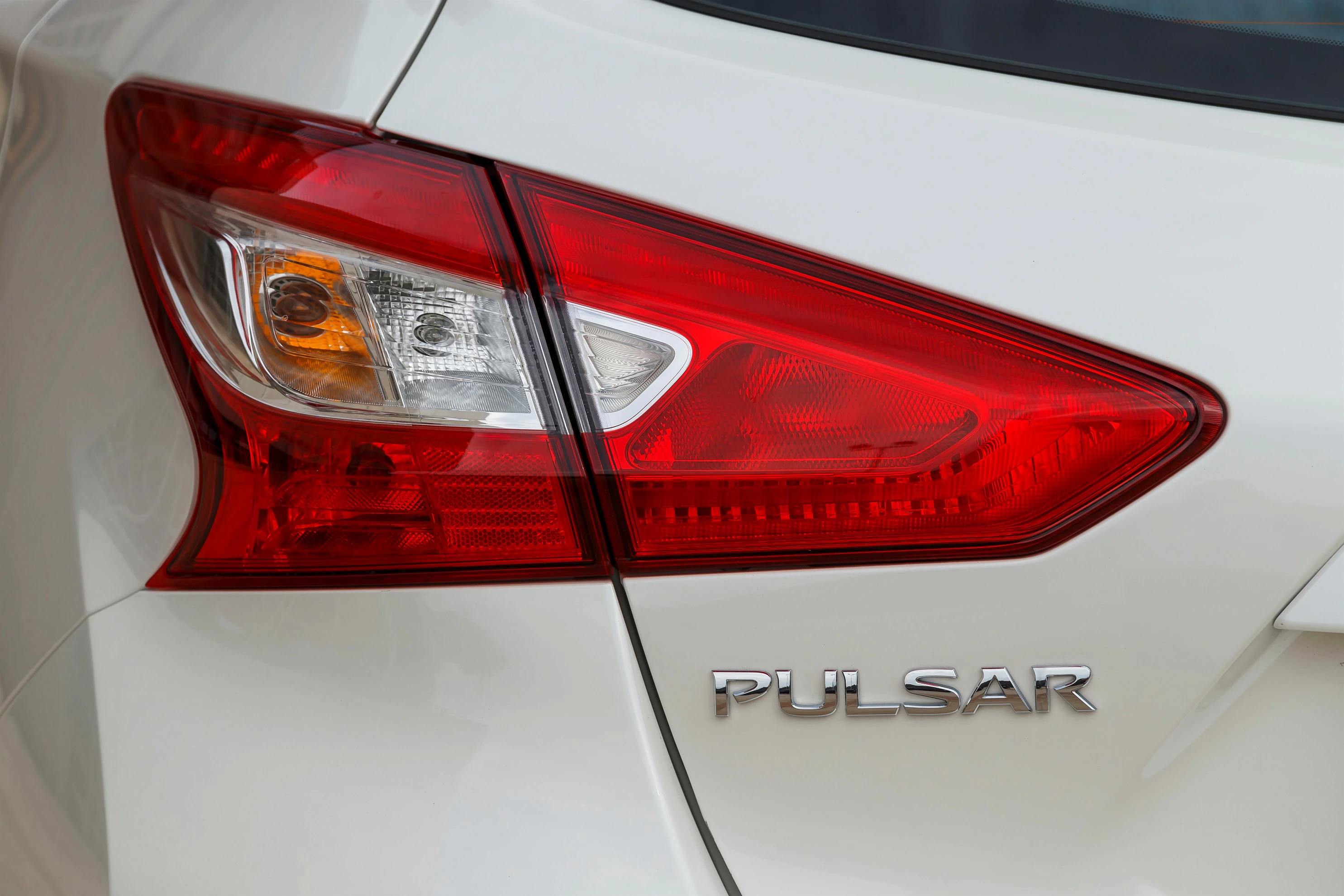 Nahaufnahme eines weißen Autos mit dem Schriftzug "PULSAR", rotes Rücklicht und Blinker auf der linken Seite sichtbar.