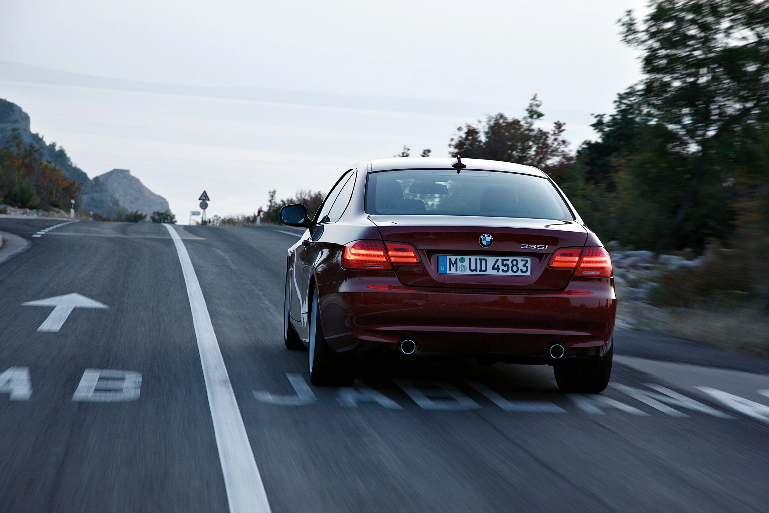 Ein roter BMW fährt auf einer Straße durch eine landschaftliche Gegend. Vor ihm liegt eine enge Kurve, umgeben von Bäumen und Hügeln.