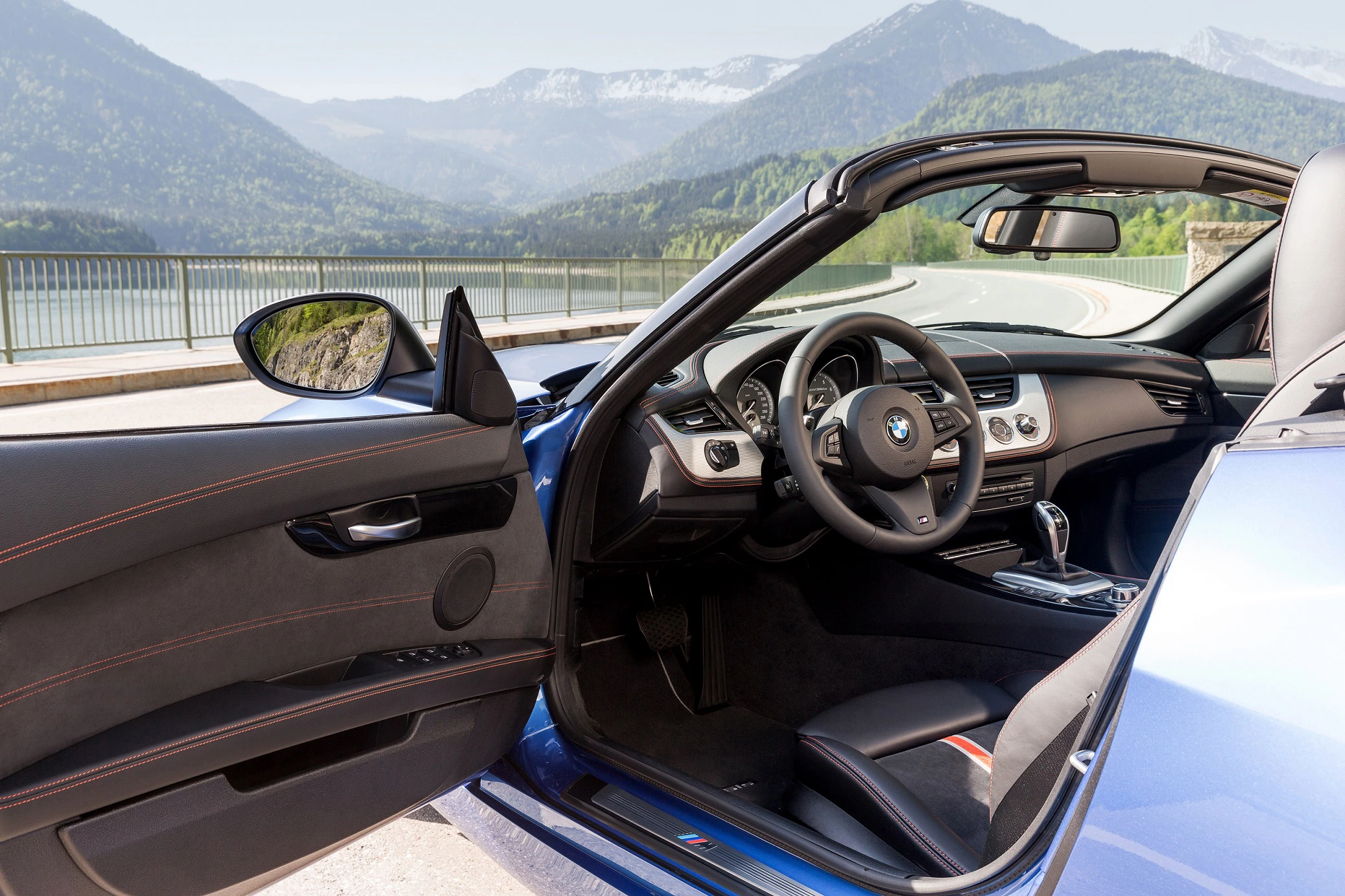 Blauer BMW Roadster mit offenem Verdeck, Blick auf das Lenkrad, vor einer malerischen Berg- und Seelandschaft geparkt.