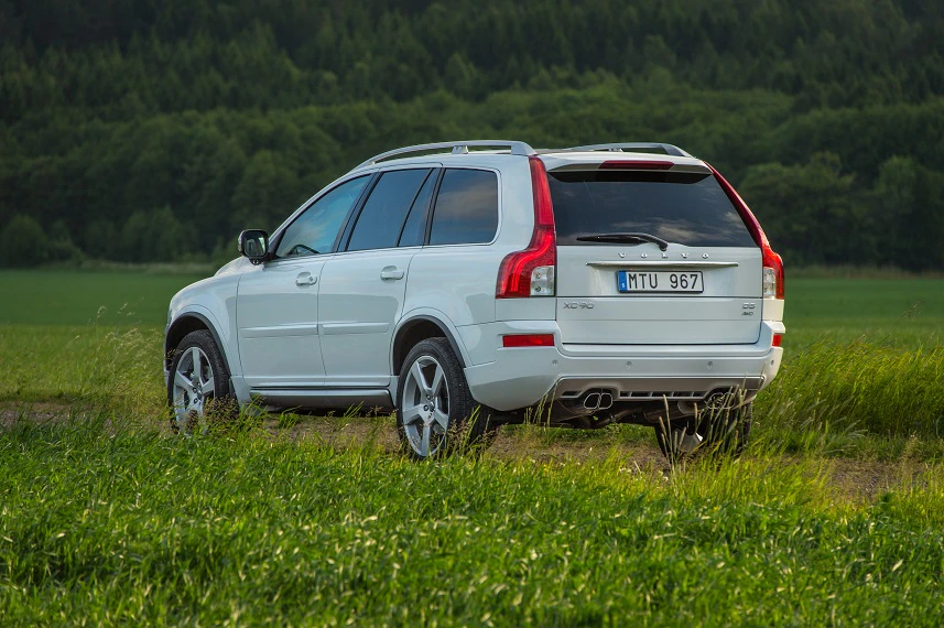Weißer SUV parkt auf einem Feldweg, umgeben von grünen Gräsern und Bäumen im Hintergrund. Links steht eine dichte Baumreihe.