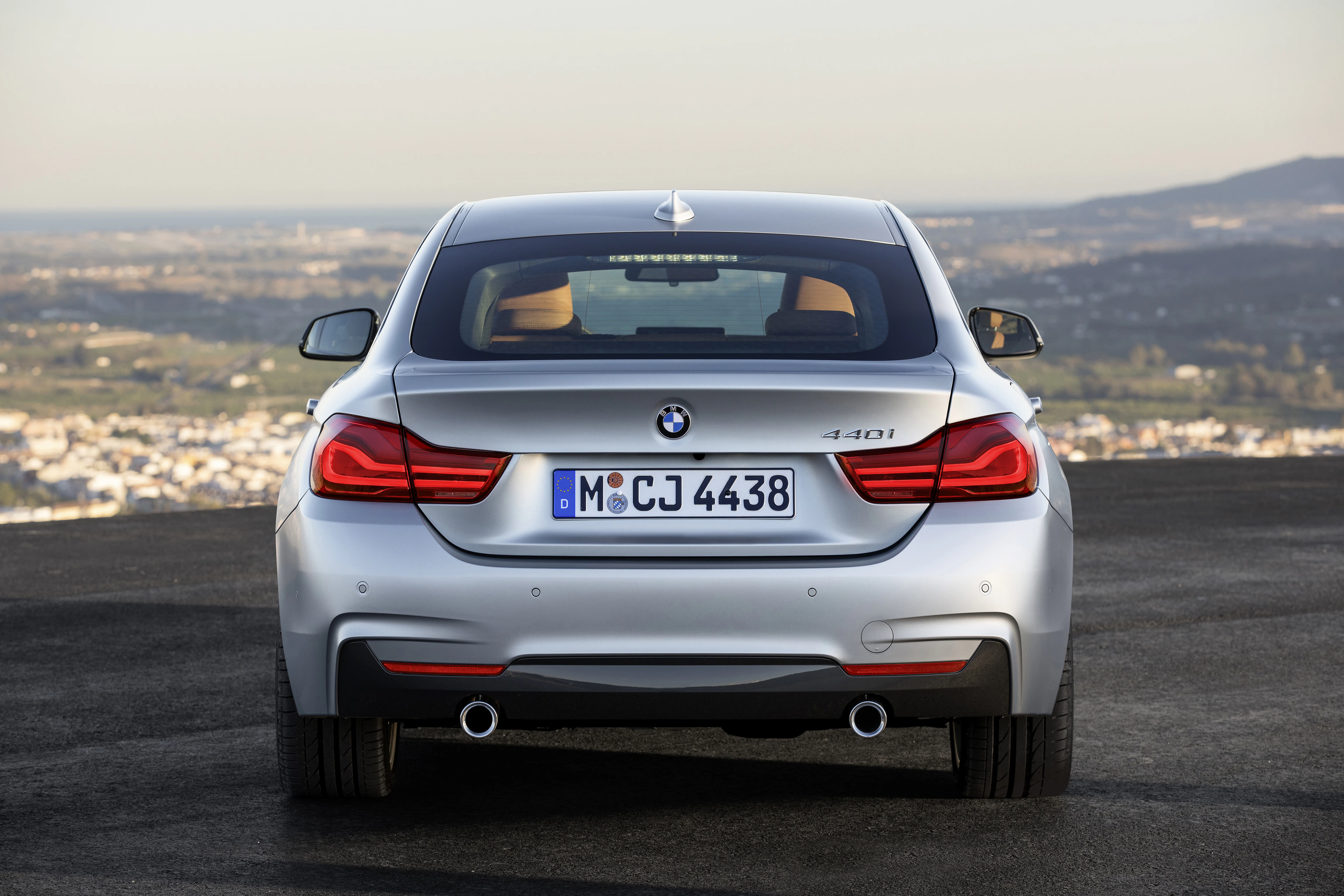 Silberner BMW 4er Gran Coupé von hinten, auf einer Anhöhe geparkt, mit Blick auf eine Stadt und Hügel im Hintergrund.