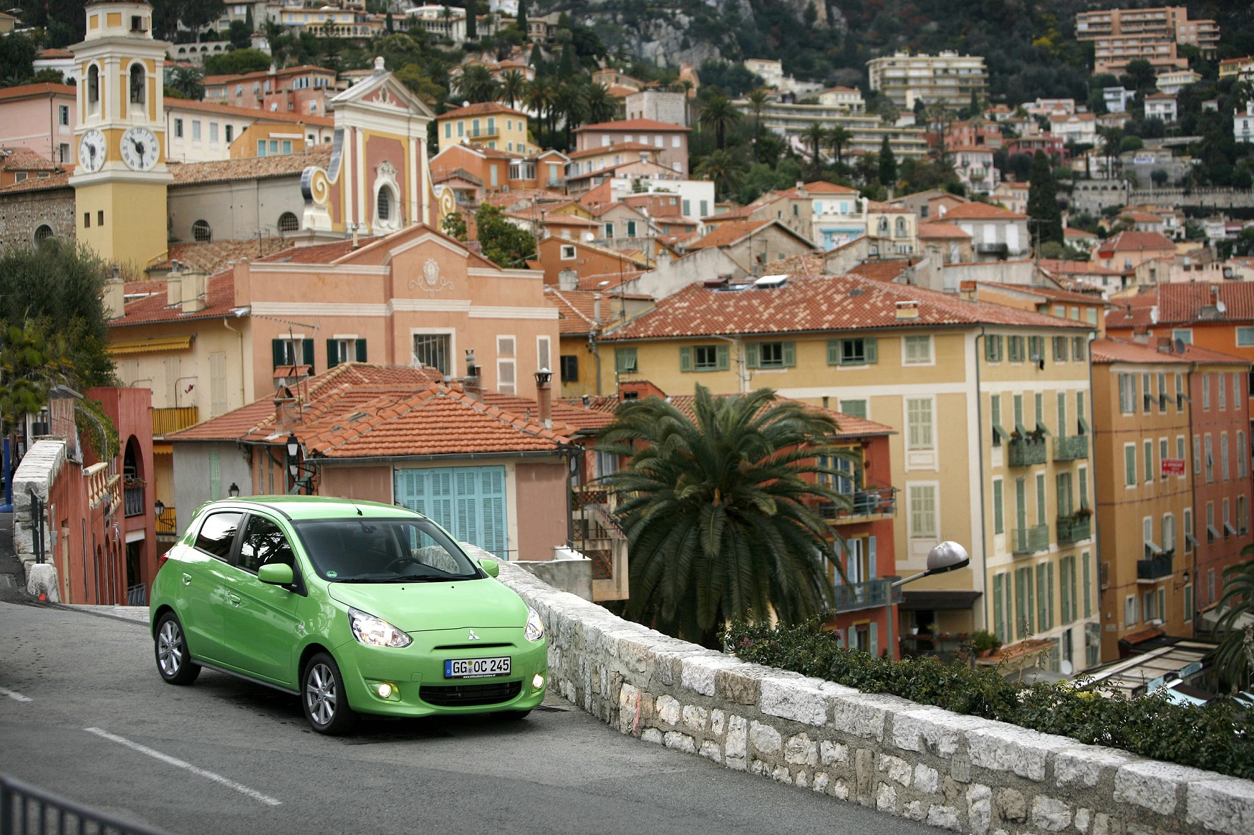 Grünes Auto auf einer Straße mit Blick auf eine malerische Stadt mit bunten Häusern und Palmen in hügeliger Umgebung.