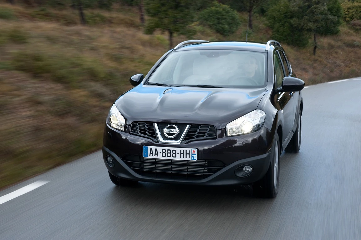 Ein schwarzer Nissan Qashqai fährt auf einer nassen Landstraße, umgeben von Bäumen und Gras.
