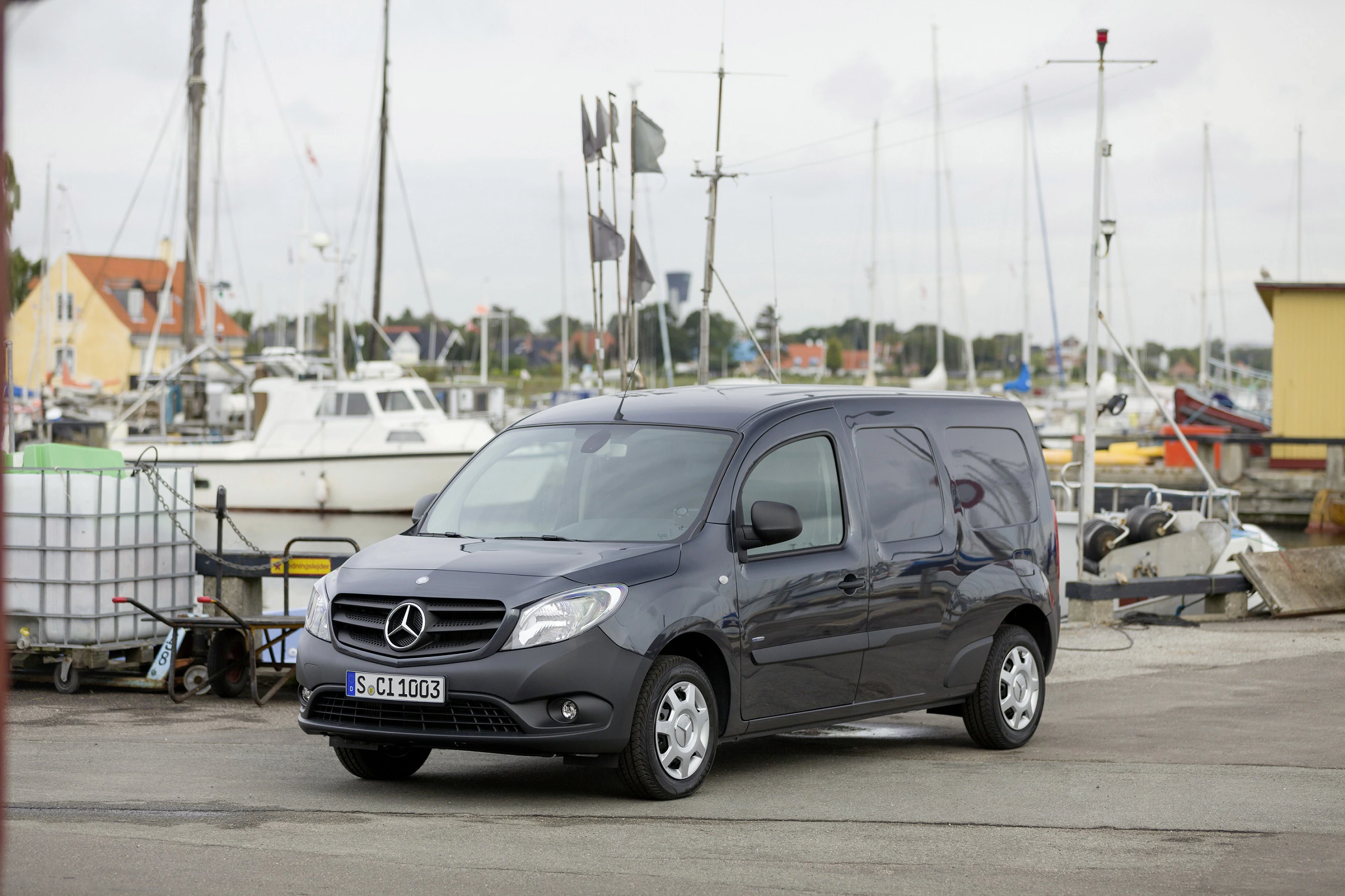 Ein grauer Mercedes-Benz Transporter parkt auf einem Hafengelände, umgeben von Segelbooten und Bootsmasten.