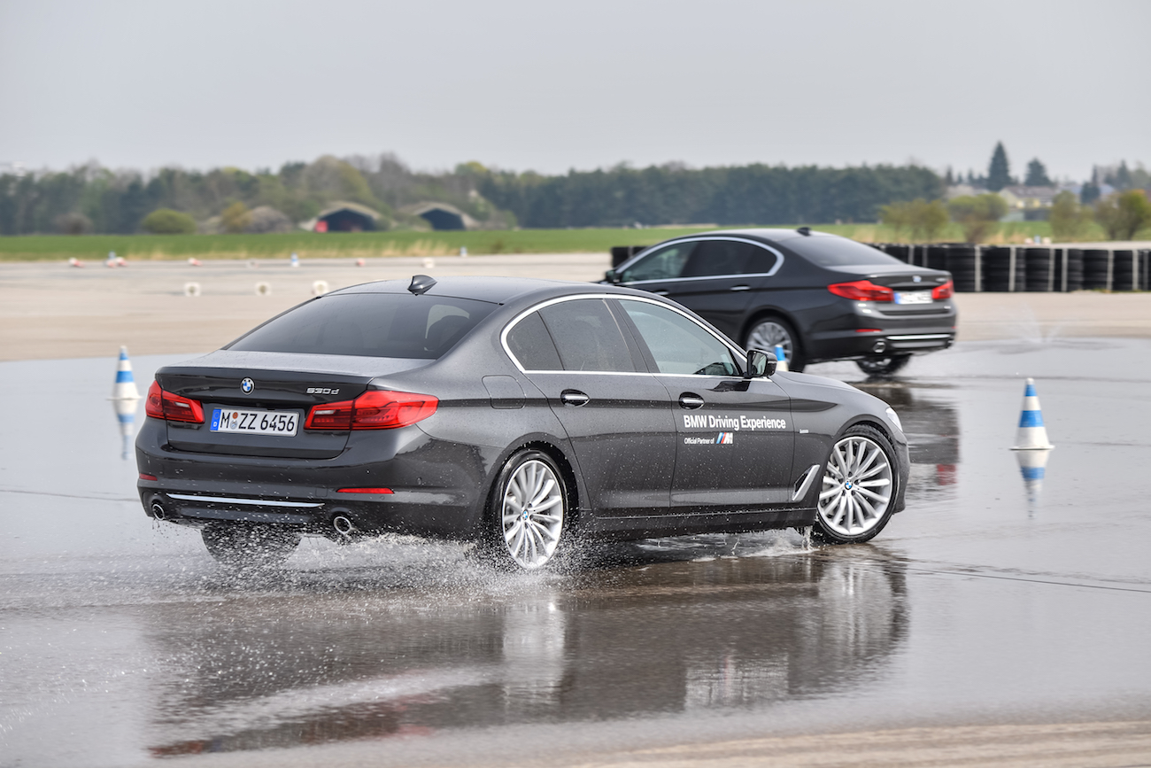 Zwei BMWs fahren in einem Fahrerlebnis-Training über eine nasse Strecke. Einer driftet leicht, im Hintergrund sind Reifenstapel sichtbar.