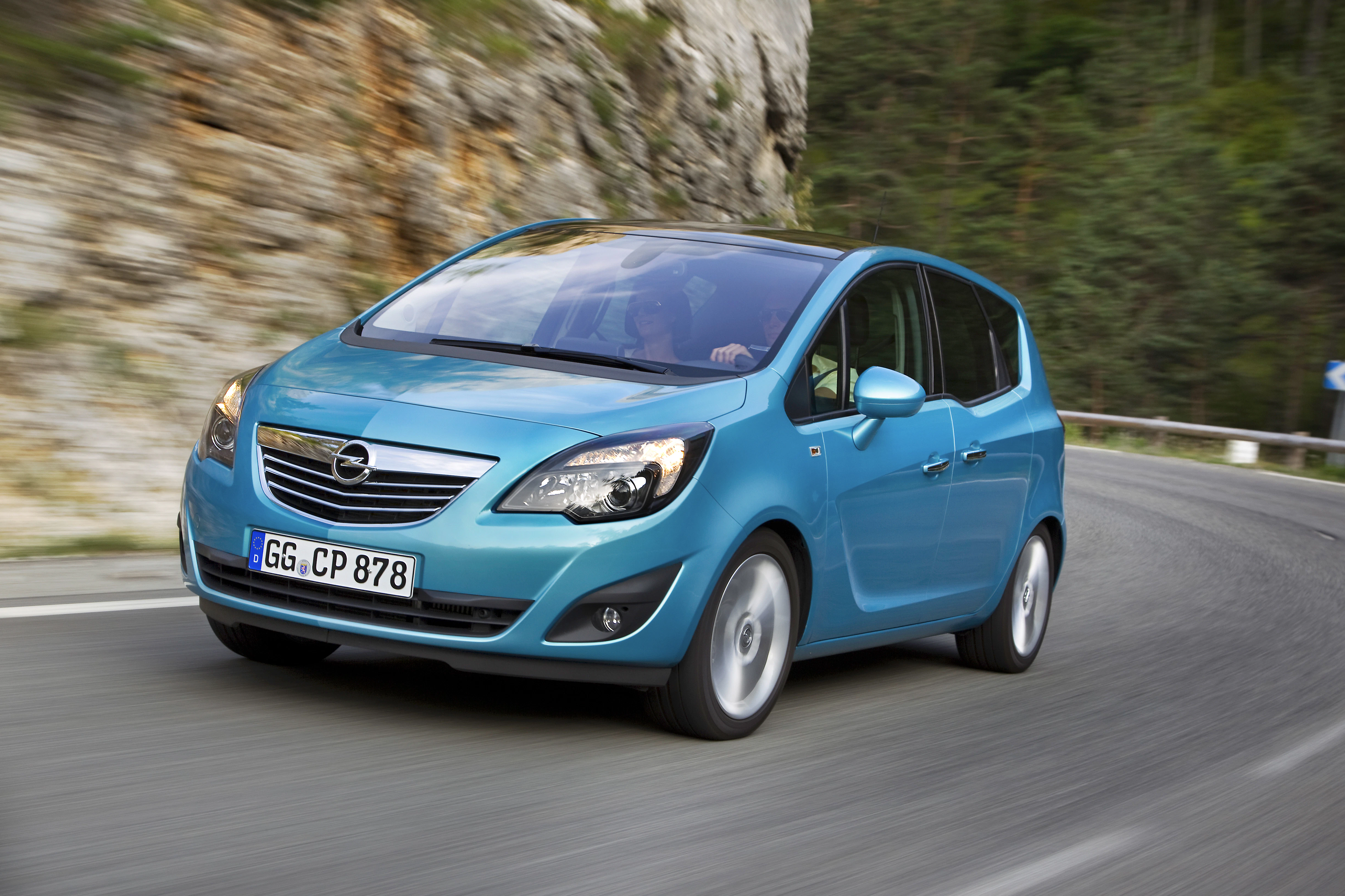 Blauer Opel Meriva fährt auf einer kurvigen Bergstraße. Im Hintergrund sind Felsen und Bäume zu sehen. Autokennzeichen sichtbar.
