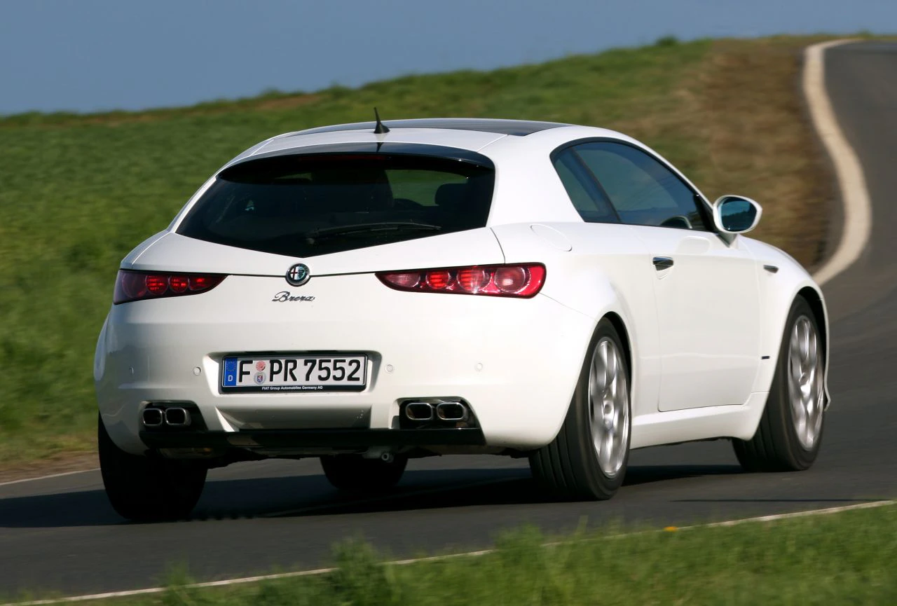 Weißer Alfa Romeo Brera fährt auf einer kurvigen Landstraße entlang grüner Felder.