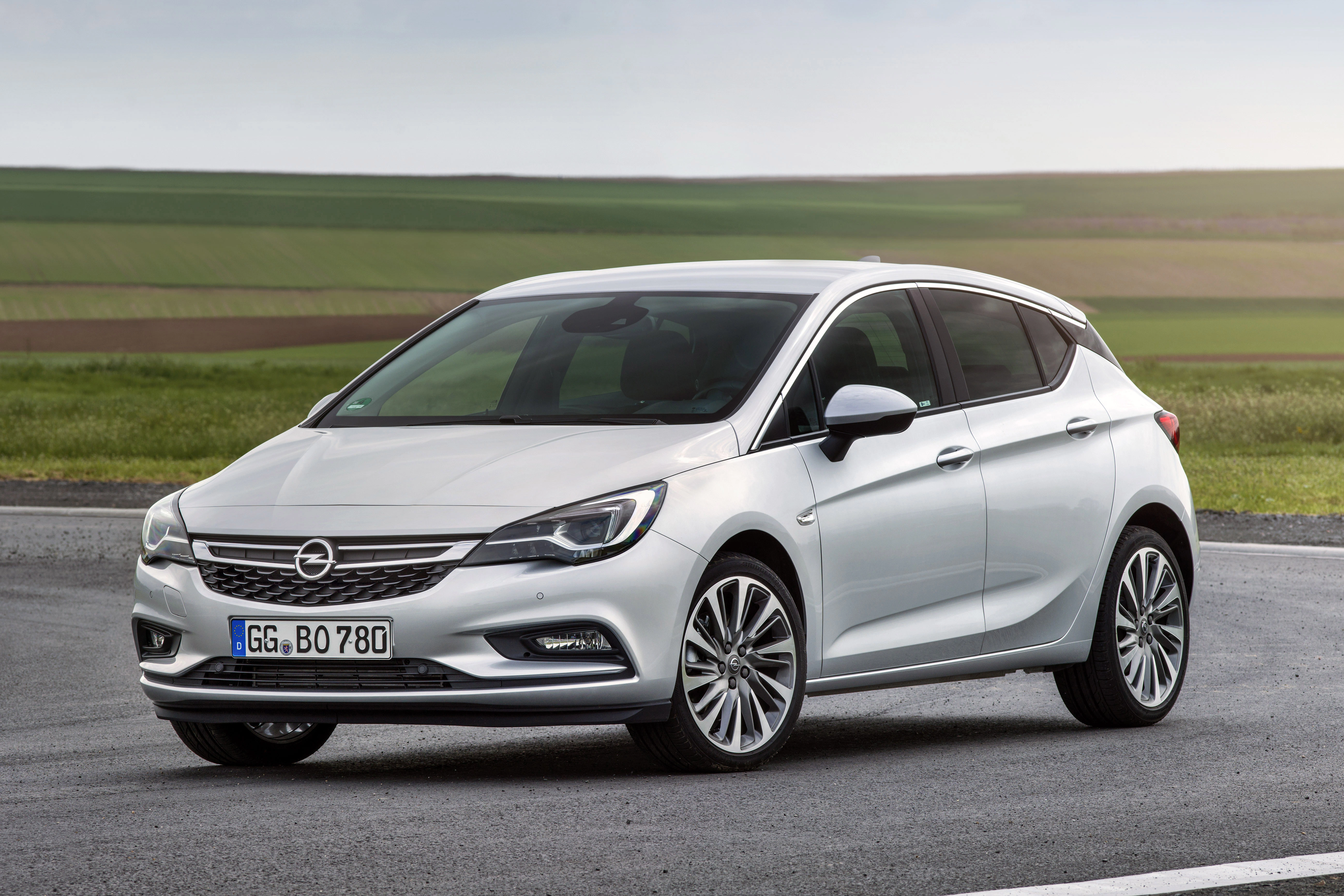 Silberner Opel Astra parkt auf einer Straße mit grünen Feldern im Hintergrund.