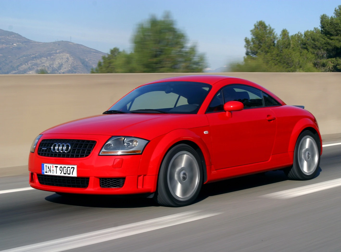 Roter Audi TT fährt auf einer Straße, im Hintergrund verschwommene Landschaft mit Bäumen und Bergen bei sonnigem Wetter.