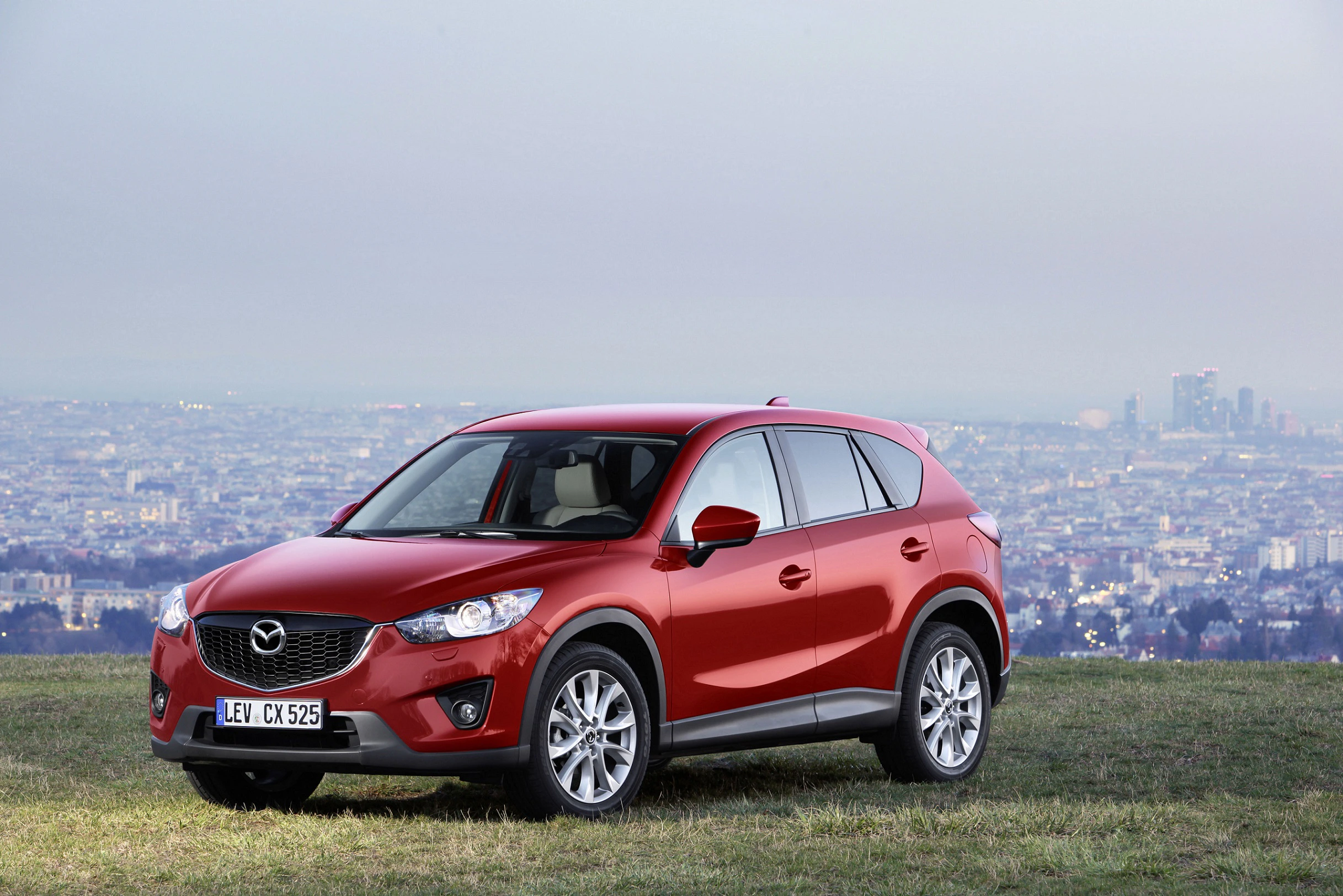 Roter Mazda CX-5 SUV parkt auf einer Wiese mit städtischem Hintergrund am Horizont unter grauem Himmel.