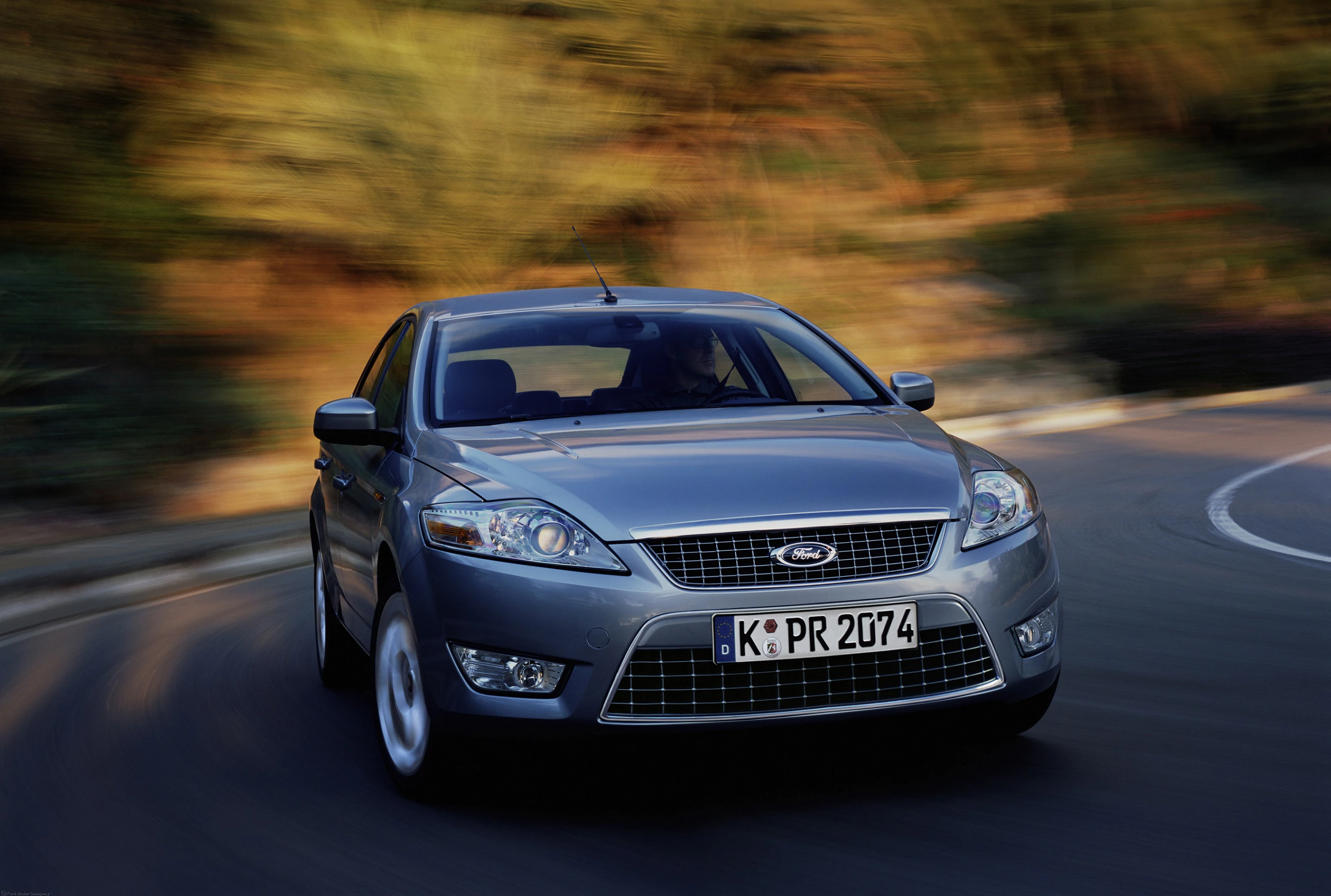 Silberner Ford Mondeo fährt dynamisch auf einer kurvigen Straße, umgeben von verschwommenem Hintergrund.