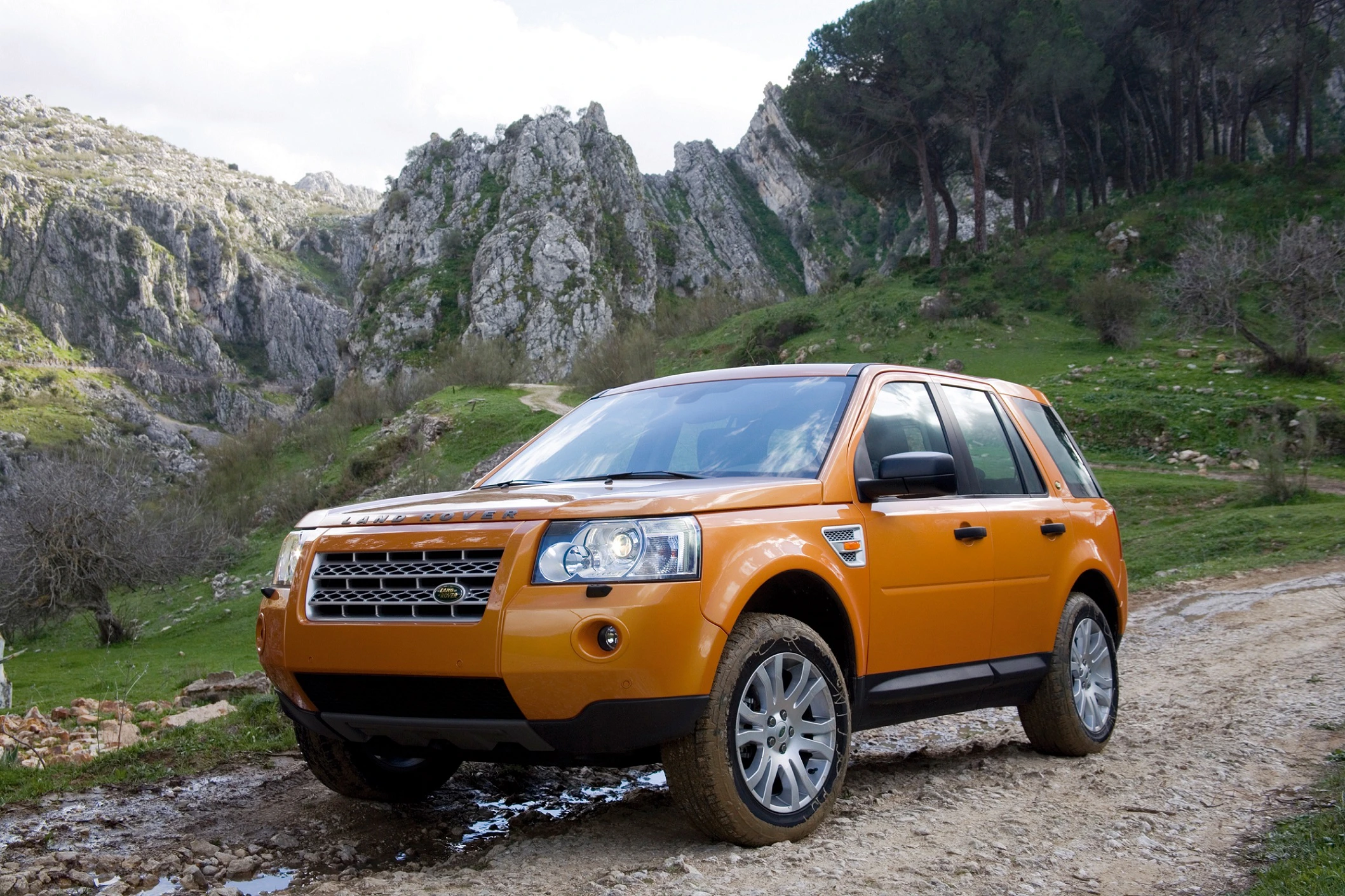 Ein orangefarbener SUV fährt auf einem felsigen Weg in einer bergigen Landschaft mit grünen Bäumen und steilen Klippen im Hintergrund.