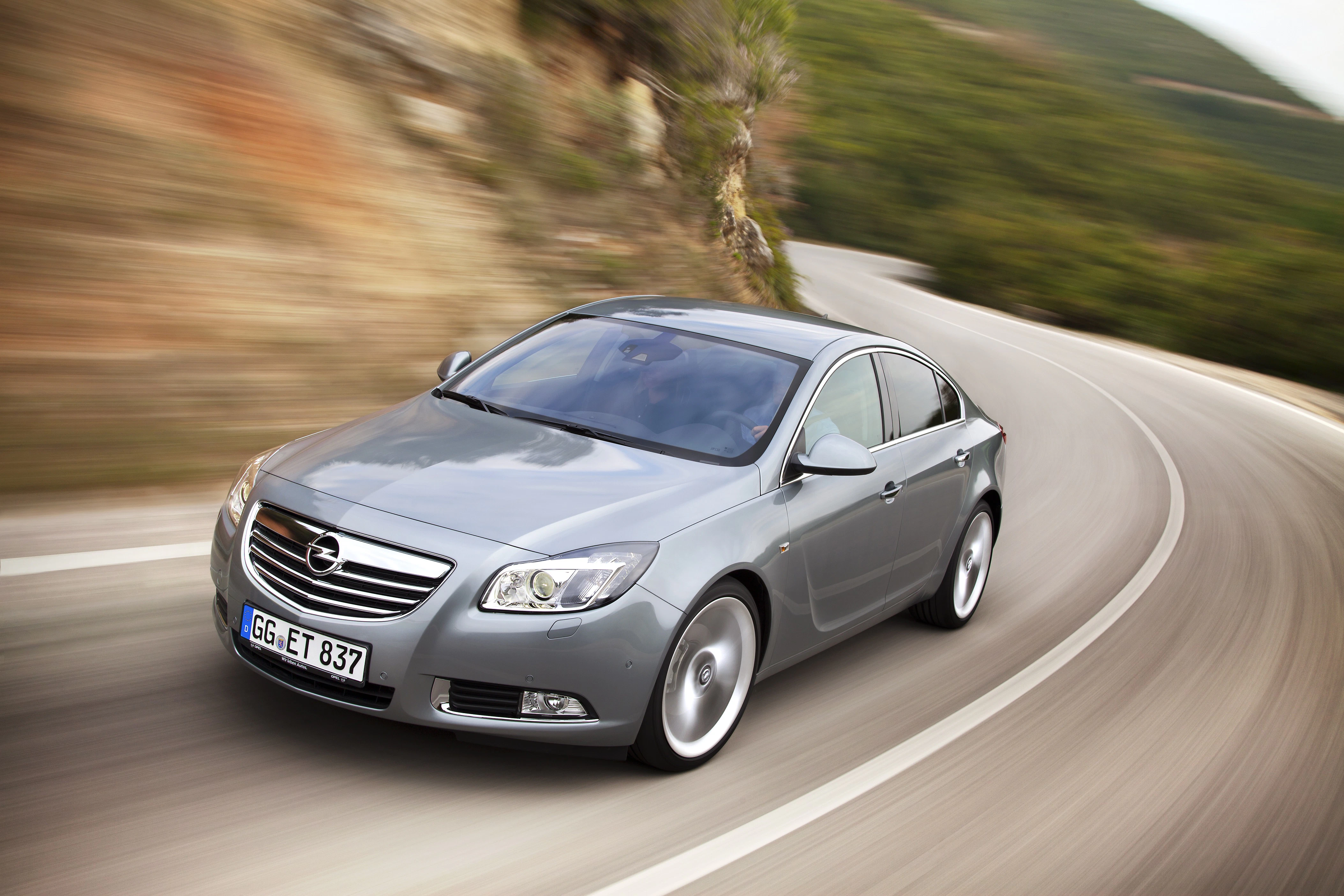 Silberner Opel fährt dynamisch auf kurvenreicher Bergstraße. Die Umgebung ist verschwommen und betont die Bewegung des Fahrzeugs.