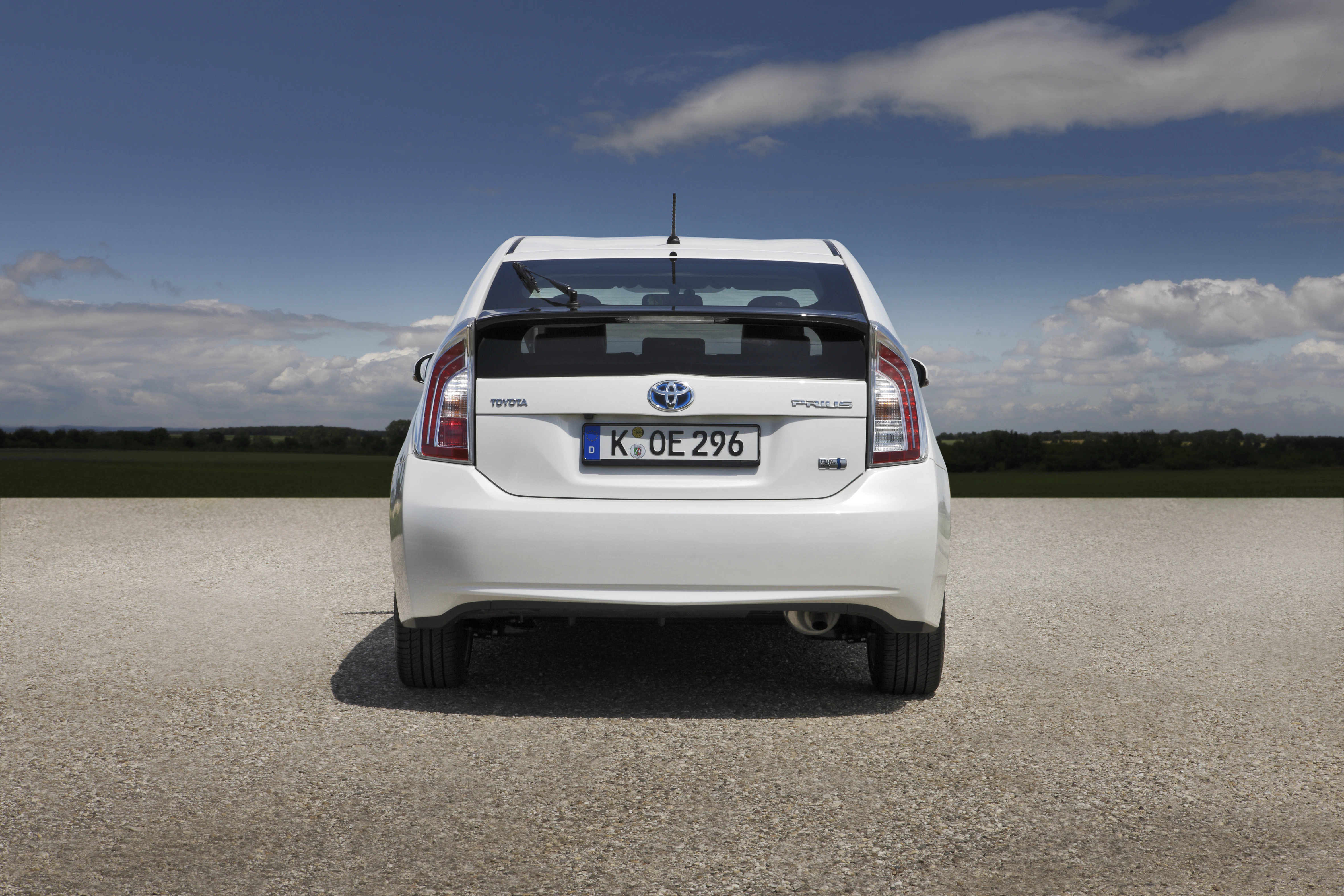 Rückansicht eines weißen Toyota Prius auf einer Straße mit einem Kennzeichen. Im Hintergrund blauer Himmel und grüne Landschaft.