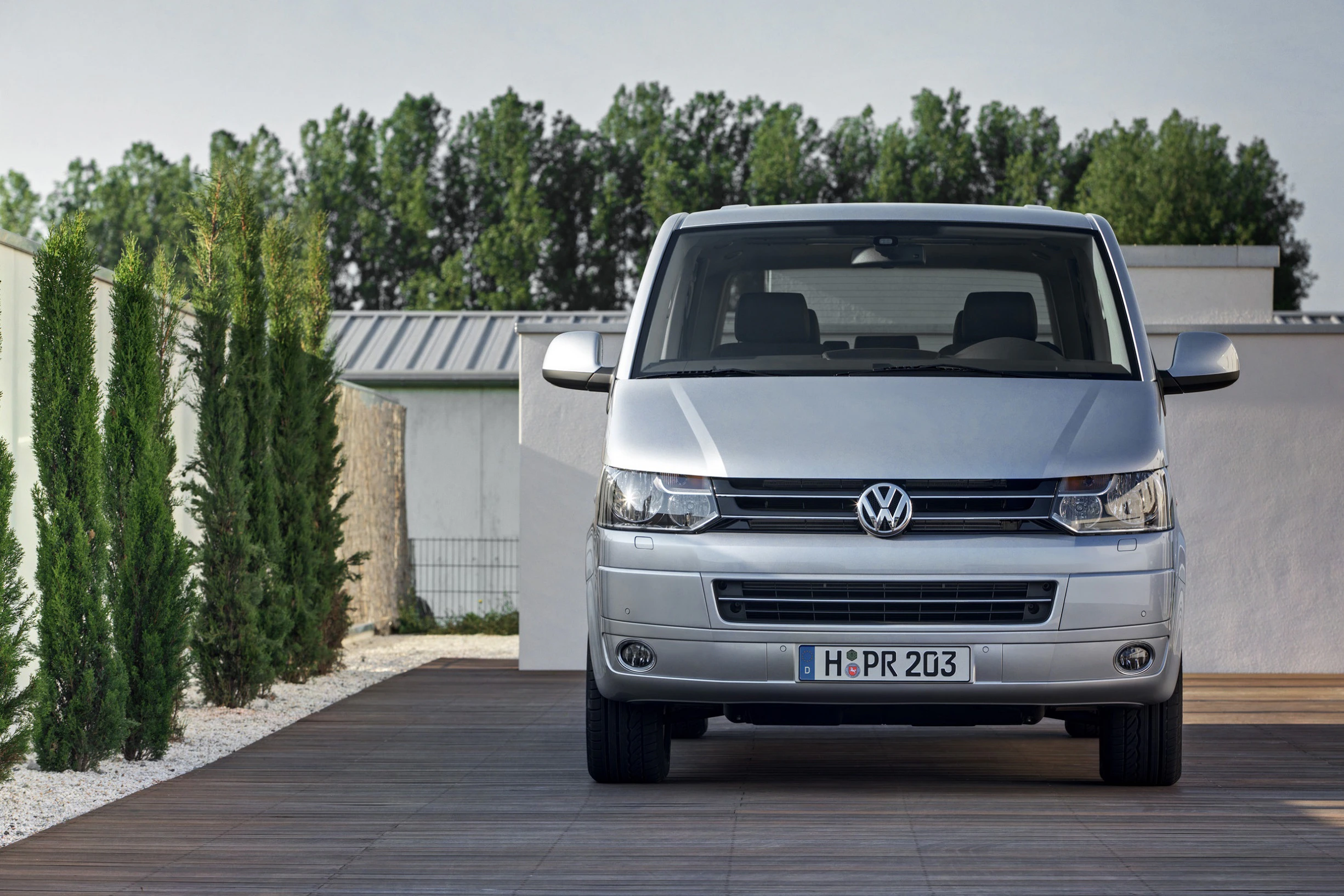 Silberner Volkswagen Transporter steht auf Holzterrasse, umgeben von grünen Pflanzen und weißem Gebäude, im Hintergrund sind Bäume zu sehen.