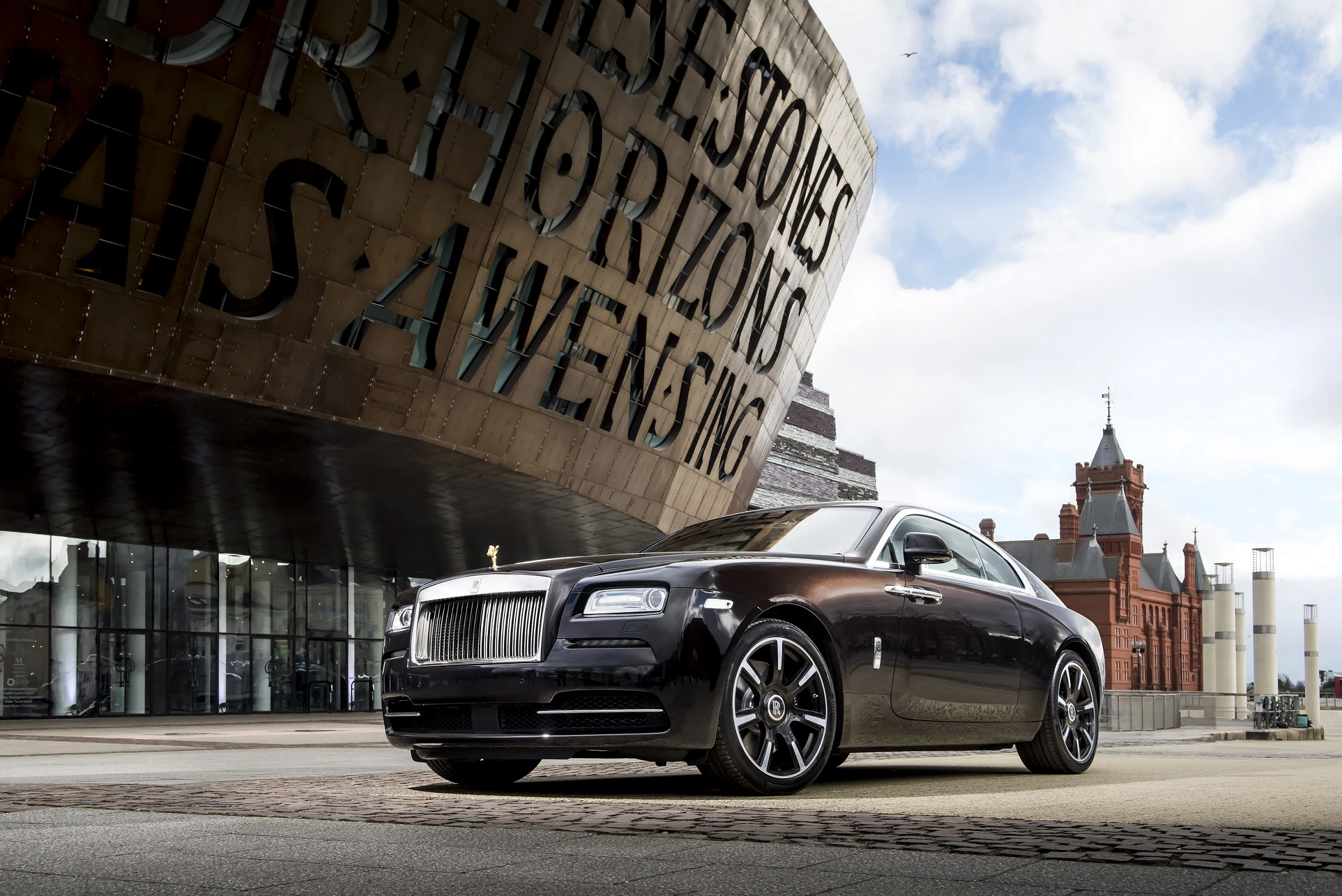 Luxuriöser schwarzer Rolls-Royce vor modernem Gebäude mit auffälliger Beschriftung und historischer Architektur im Hintergrund.