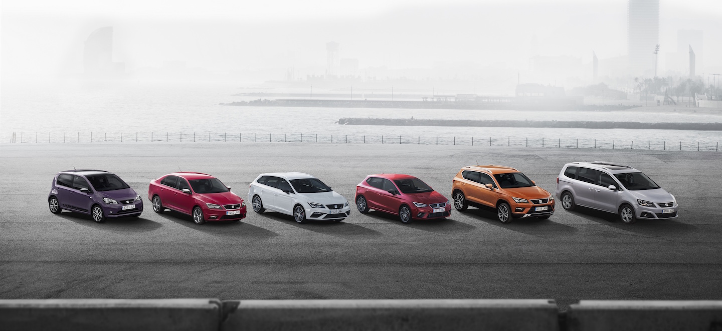 Sechs bunte Autos stehen nebeneinander auf einer Promenade mit verschwommenem Hintergrund und Wasser.