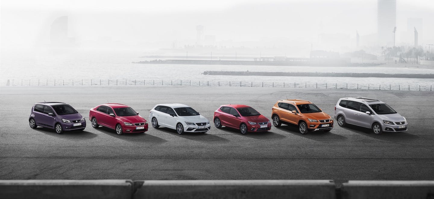 Sechs bunte Autos stehen nebeneinander auf einer Promenade mit verschwommenem Hintergrund und Wasser.