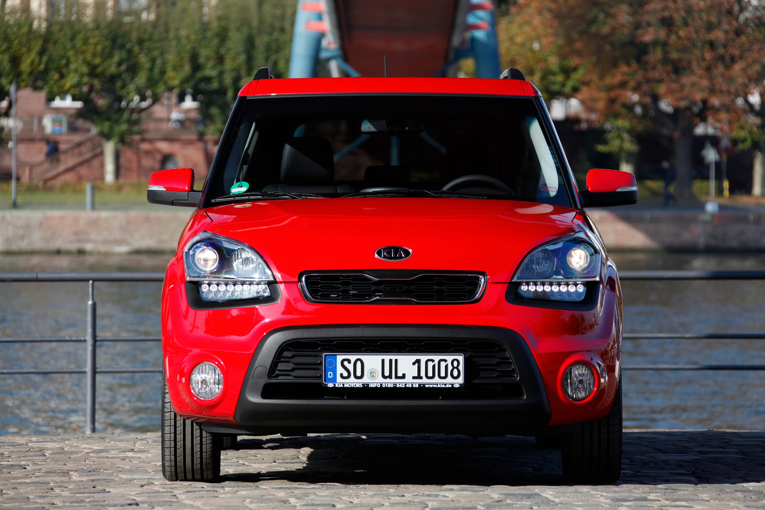 Roter Kia Soul steht auf Kopfsteinpflaster vor einem Flussufer mit Brücke und Bäumen im Hintergrund.
