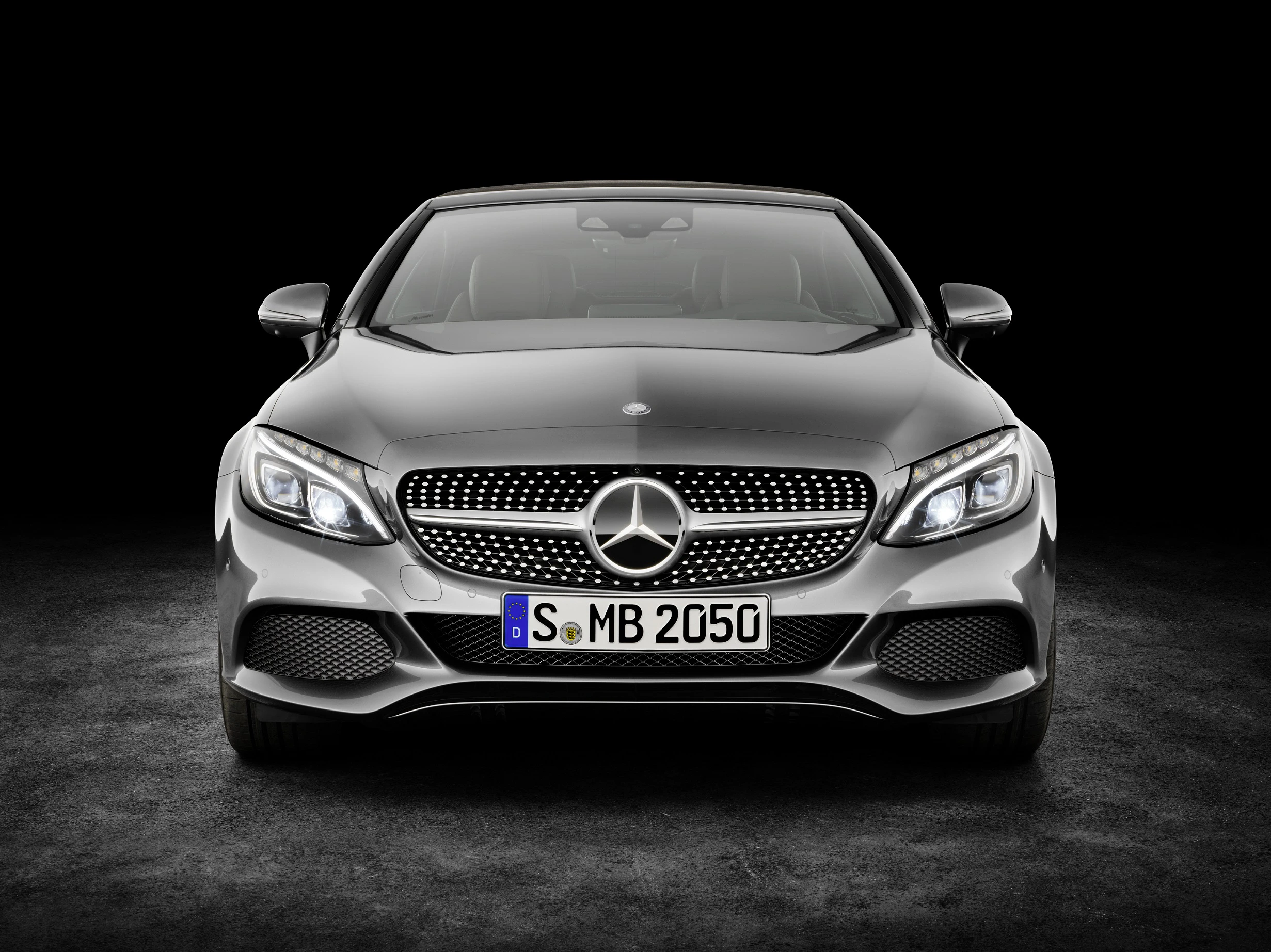 Grauer Mercedes-Benz von vorne vor schwarzem Hintergrund, mit leuchtenden Scheinwerfern und sichtbarem Markenlogo auf dem Kühlergrill.