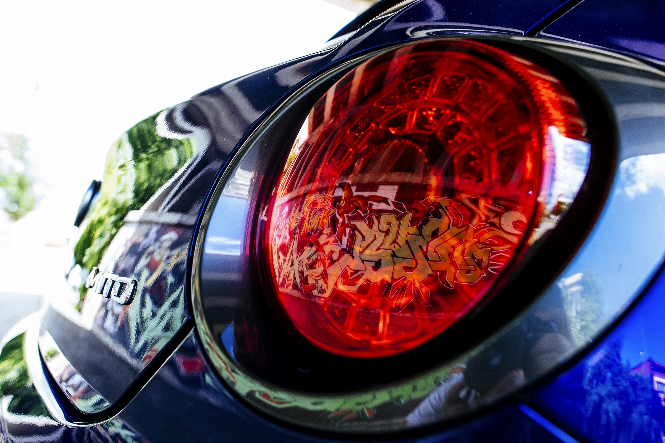 Nahaufnahme eines roten Rücklichts eines blauen Autos, das im Sonnenlicht leuchtet. Reflektionen und Graffiti-Muster auf der Oberfläche sichtbar.