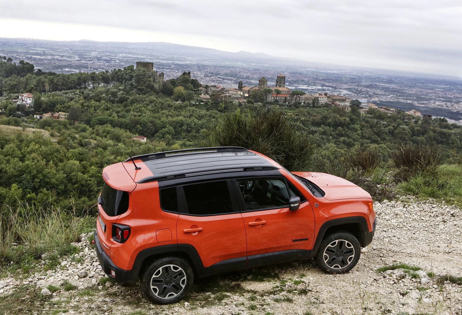 Ein rotes Auto fährt einen felsigen Hügel hinunter mit Blick auf eine bewaldete Landschaft und eine historische Stadt im Hintergrund.