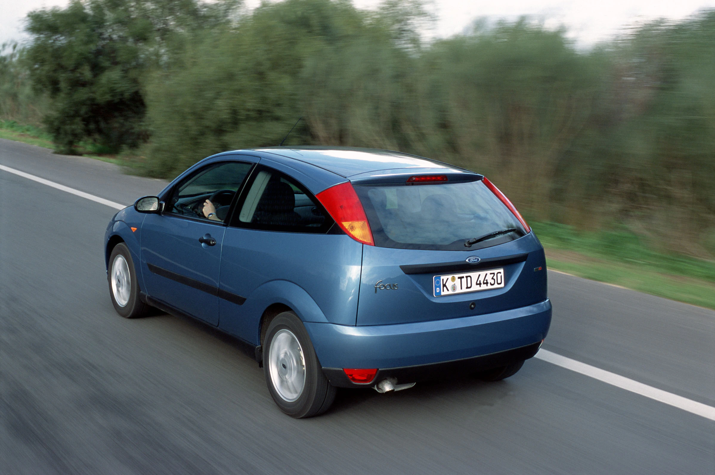 Blauer Ford Focus fährt auf einer Landstraße, umgeben von unscharfer Natur. Fahrzeug zeigt Heckansicht, keine anderen Autos in Sicht.