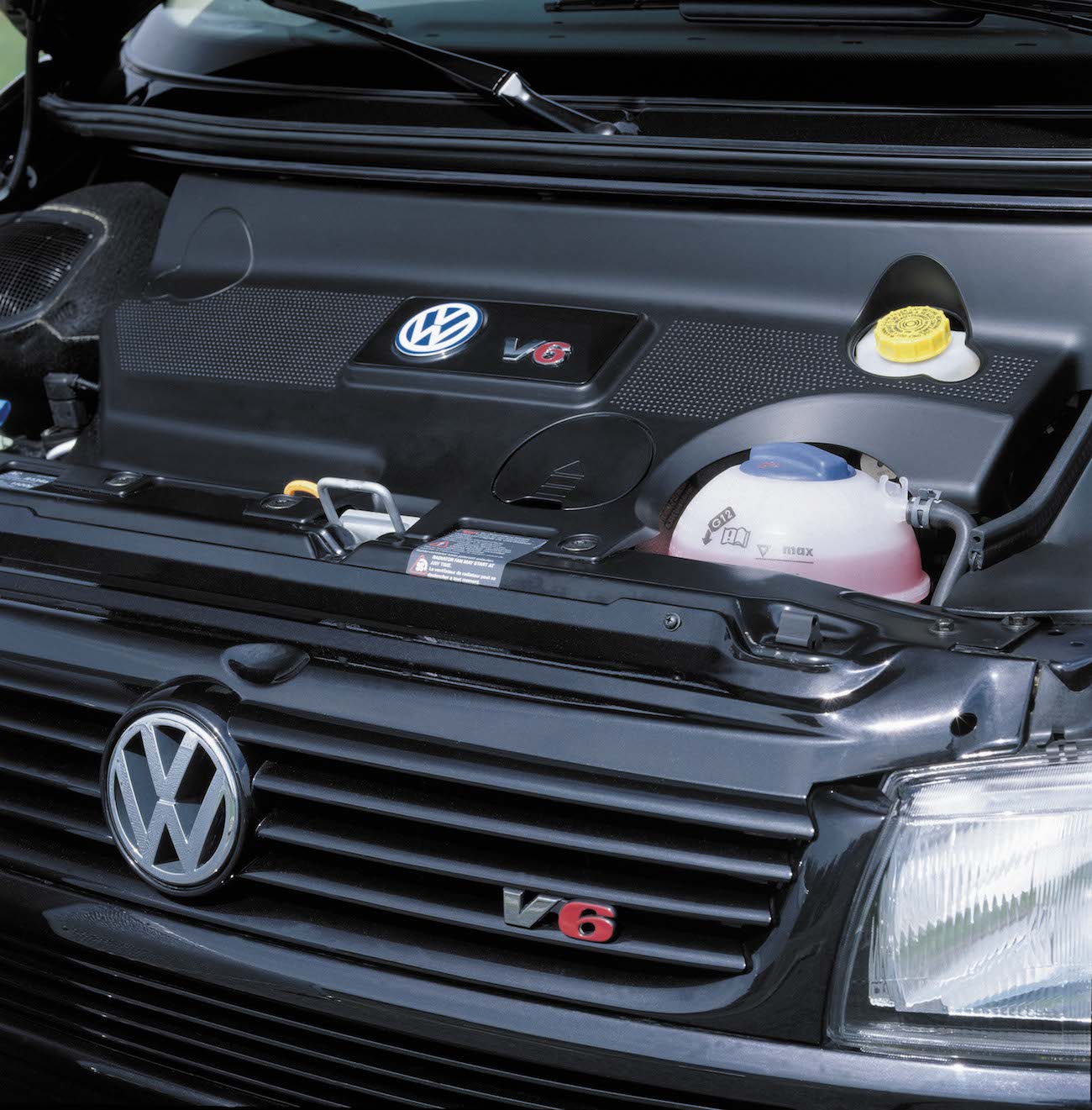 Motorraum eines Volkswagen mit V6-Motor. Sichtbare Elemente sind das Kühlergrill-Emblem, ein Kühlmittelbehälter und diverse Abdeckungen.