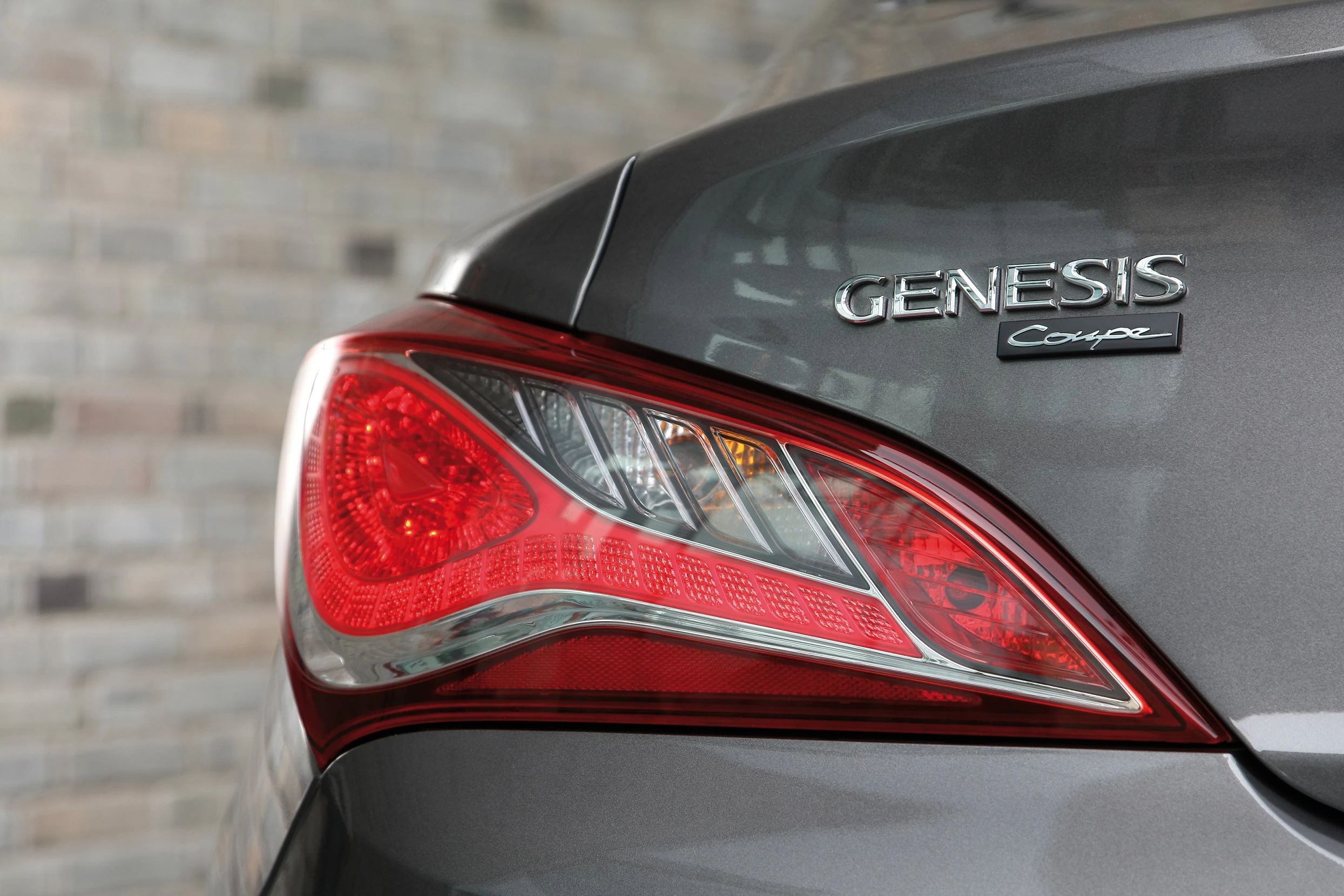 Nahaufnahme eines grauen Genesis Coupé-Heck, zeigt Rücklicht und Emblem. Hintergrund aus Backsteinmuster.