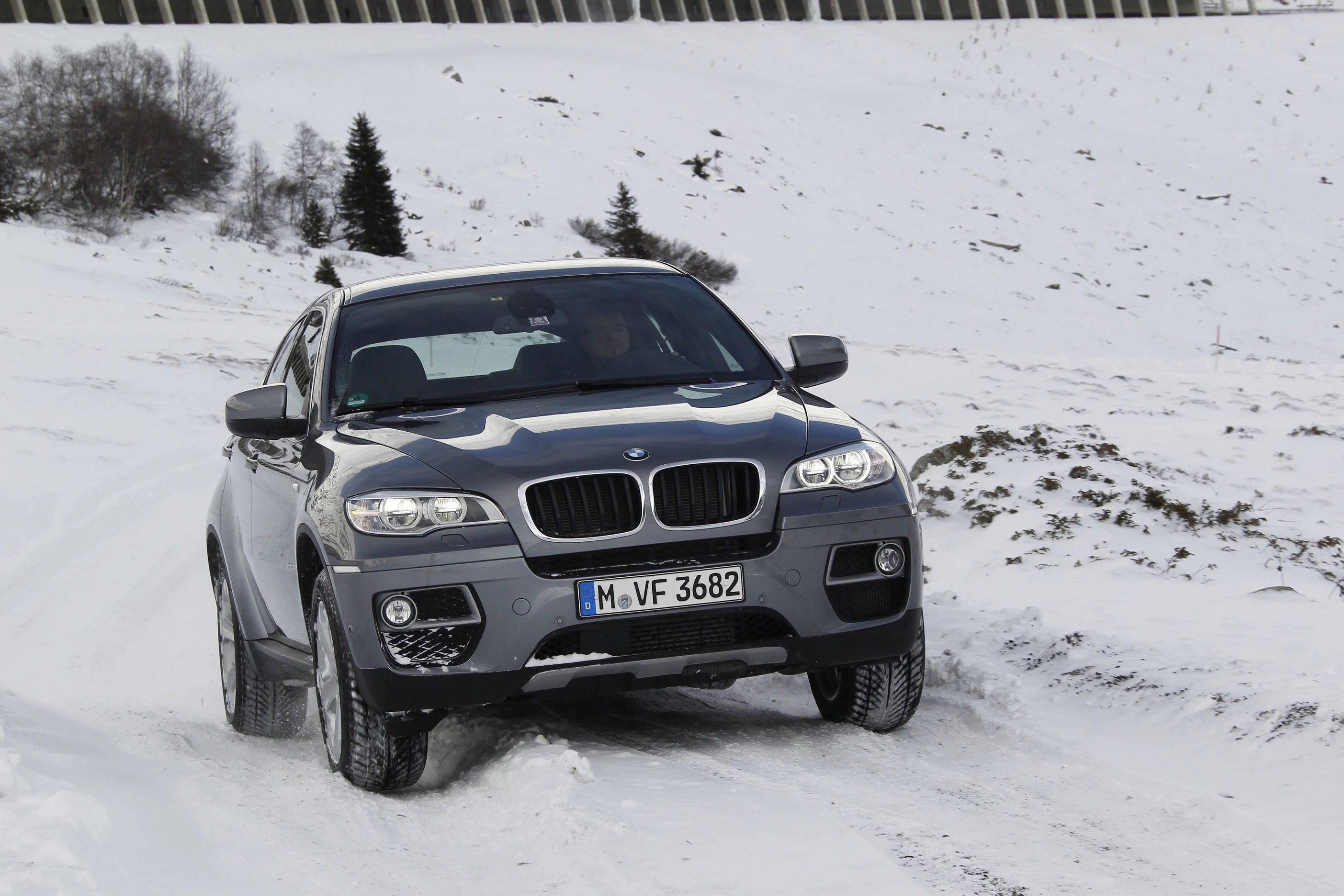 Ein schwarzer BMW X6 fährt auf einer schneebedeckten, kurvigen Straße; im Hintergrund sind Bäume und eine Mauer sichtbar.