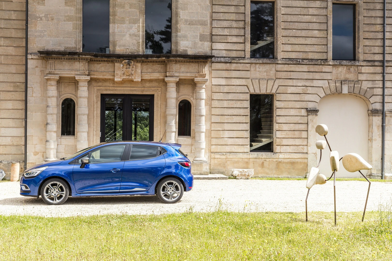Blaues Auto vor einem historischen Gebäude mit Steinskulpturen im Vordergrund auf einer Rasenfläche.