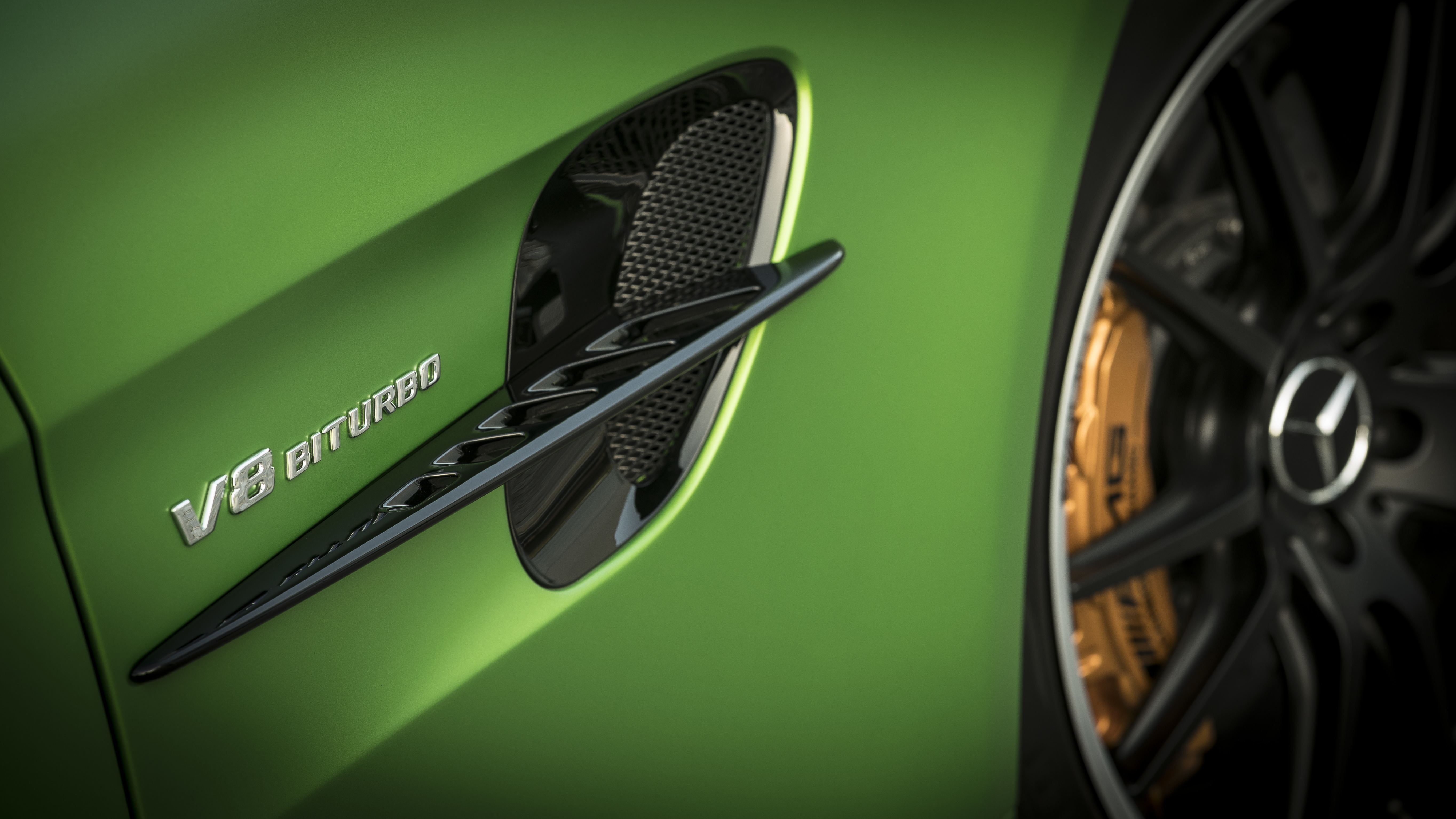 Nahaufnahme eines grünen Autos mit "V8 Biturbo"-Plakette und einem teilweise sichtbaren schwarzen Rad mit goldenen Bremssätteln.