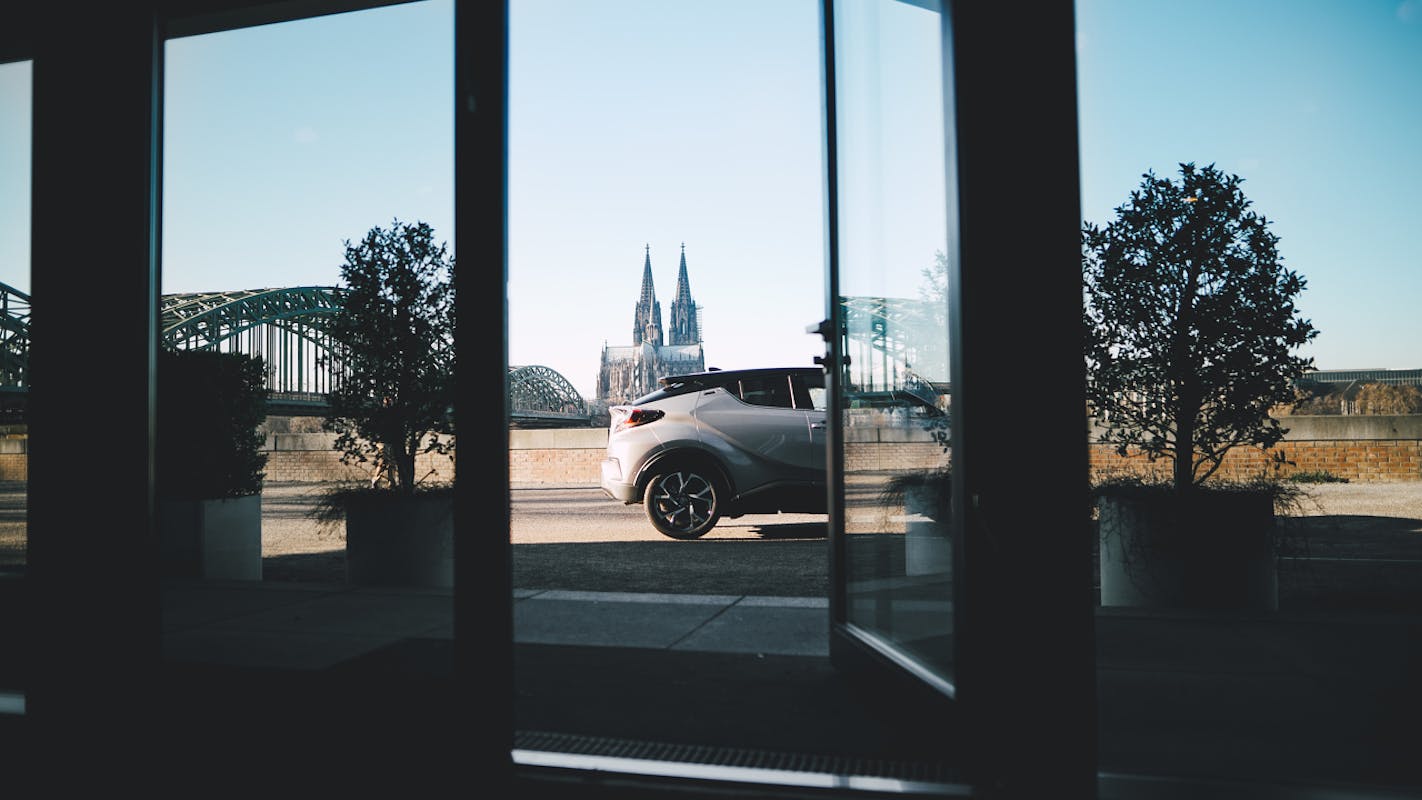 Ein weißes Auto steht vor dem Kölner Dom, eingerahmt von zwei Bäumen und einer Fensteransicht. Im Hintergrund ist die Hohenzollernbrücke zu sehen.