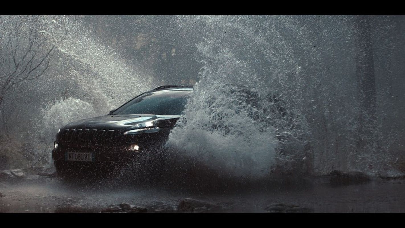 Auto fährt durch spritzendes Wasser in einer bewaldeten Umgebung bei Regen. Dynamische Aufnahme mit viel Bewegung und Wasserspritzern.
