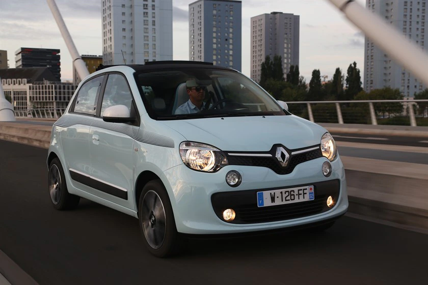 Hellblauer Renault Twingo fährt über eine Brücke, im Hintergrund moderne Hochhäuser; Person am Steuer, sonniger Tag.