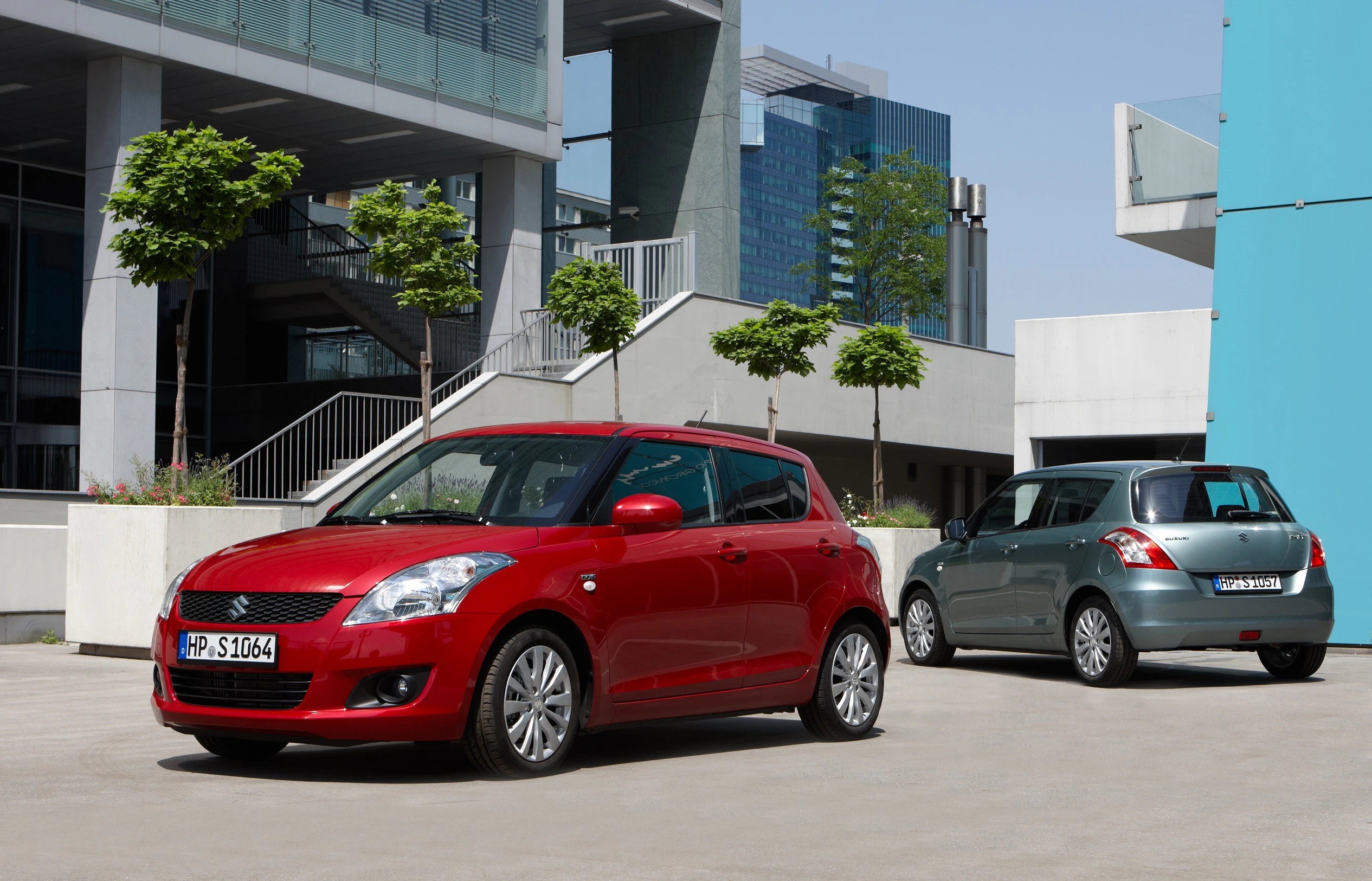 Zwei Suzuki Swift Autos parken vor modernen Bürogebäuden. Ein rotes Auto vorne, ein graues Auto im Hintergrund.