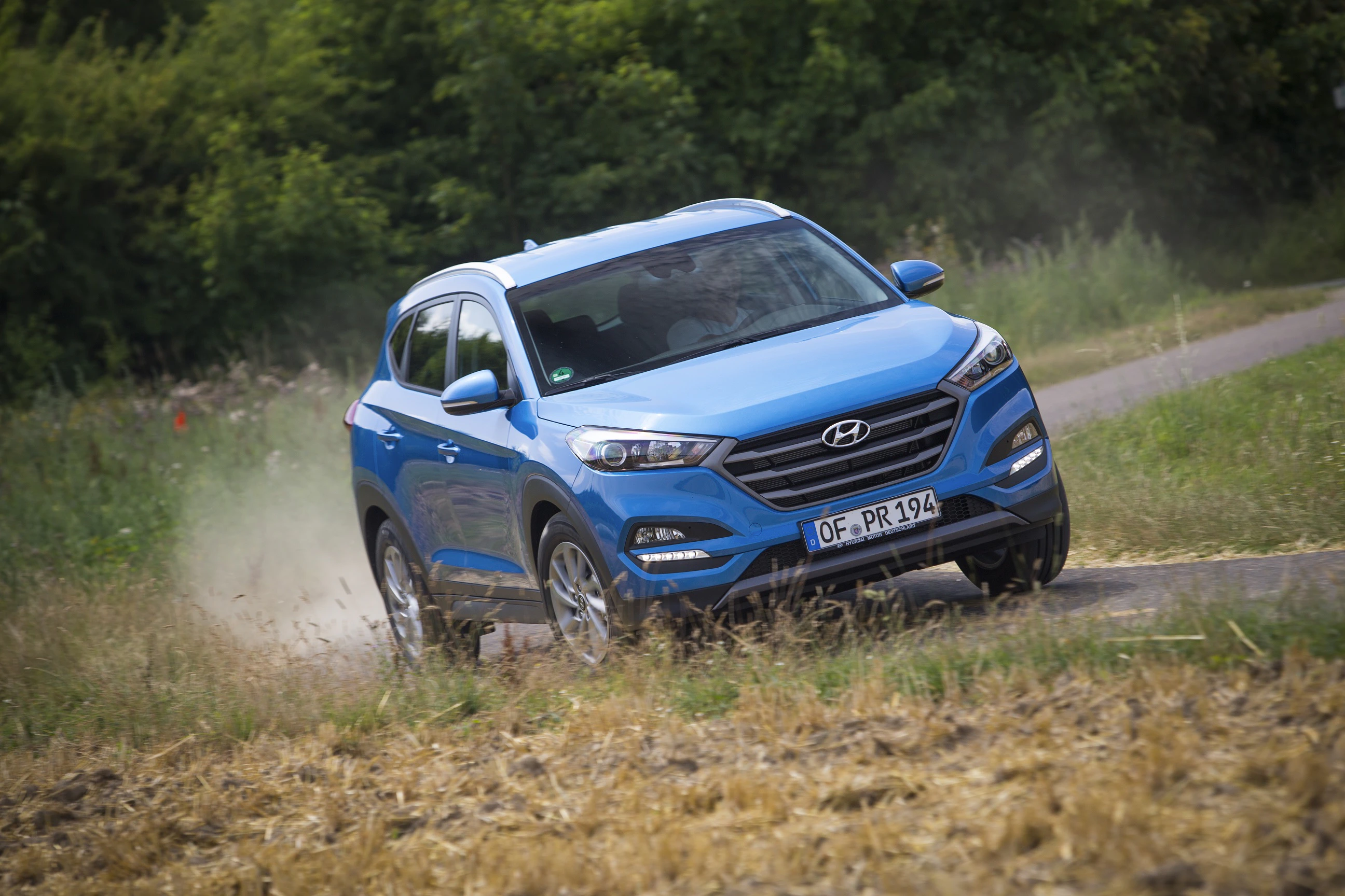 Blauer SUV fährt dynamisch auf einer ländlichen Straße, aufgewirbelter Staub, grüne Büsche im Hintergrund.