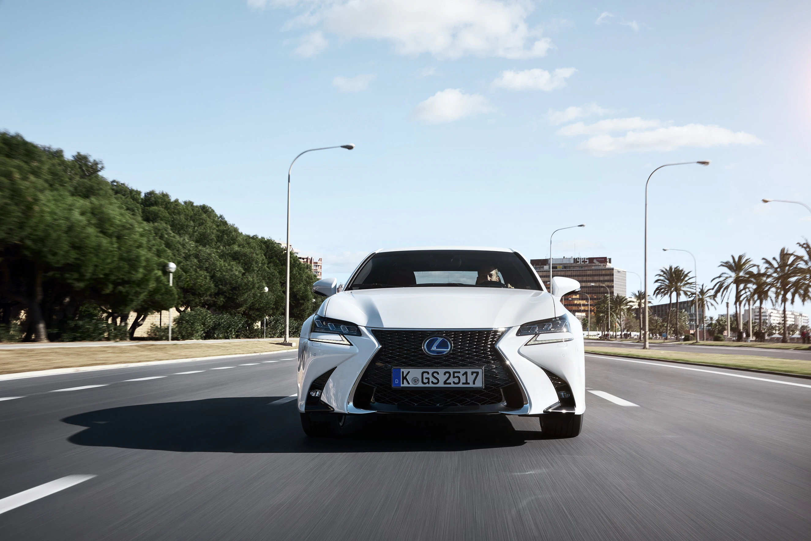 Ein weißer Lexus fährt auf einer breiten Straße mit Palmen, Bäumen und Laternen bei klarem Himmel. Nummernschild: K-GS 2517.
